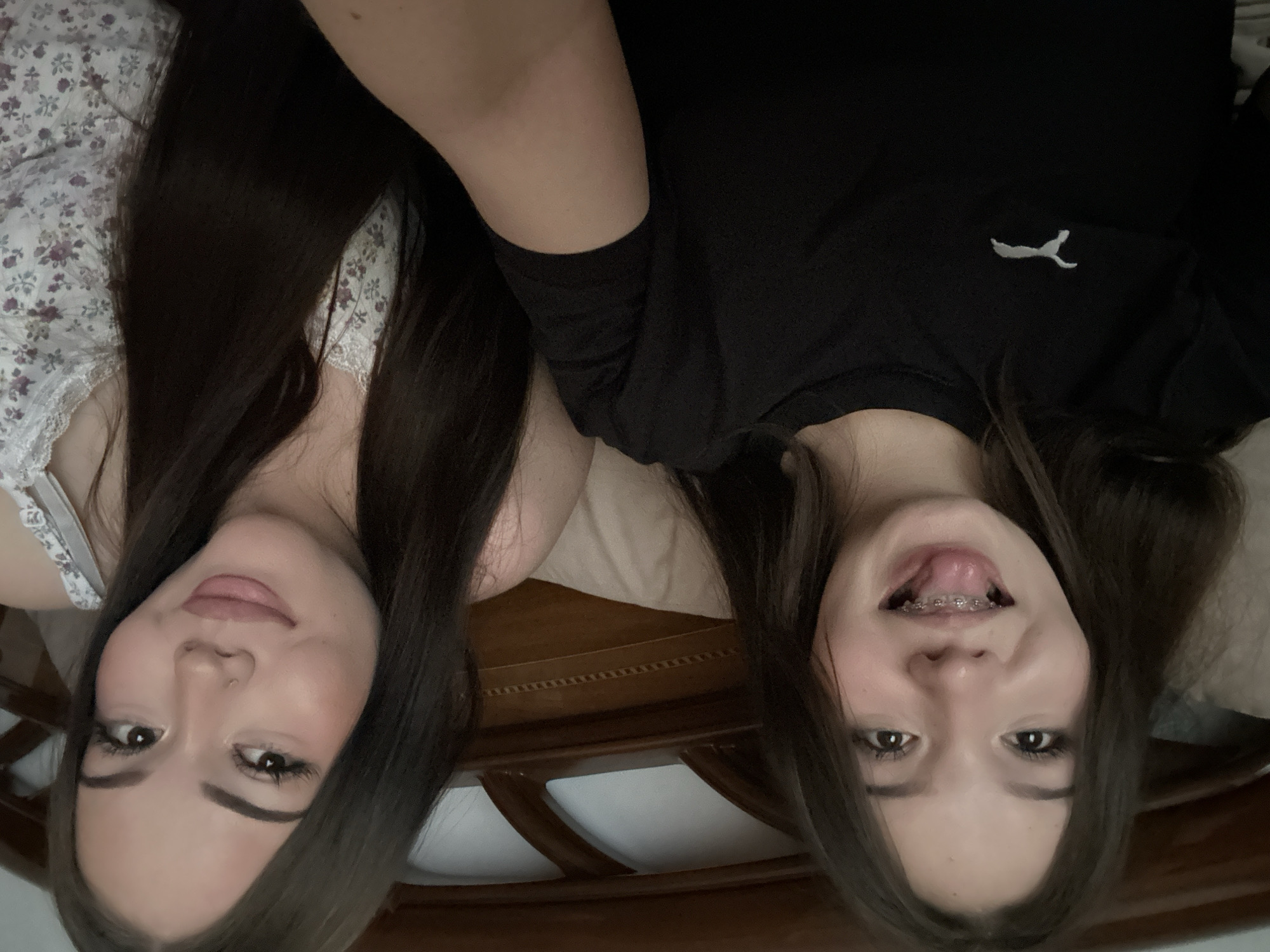 This image features two young women lying down, with their heads oriented towards the bottom of the frame, appearing upside down. They are positioned side-by-side, looking directly at the camera.
The woman on the left (viewer's left, but her right side in natural orientation) has long, straight dark brown hair. She is wearing a light-colored top with a delicate floral pattern in shades of white and purple, featuring a lace trim along the neckline. Her expression is calm, with a subtle, closed-mouth smile, and she has a small piercing on her nose.
The woman on the right (viewer's right, but her left side in natural orientation) also has long, straight dark brown hair. She is wearing a plain black t-shirt with a white Puma logo visible on the left side of her chest. Her expression is more playful; she has her mouth open, with her tongue slightly extended, revealing dental braces on her upper teeth.
They are lying on what appears to be a bed, with light-colored bedding visible. A dark brown wooden headboard or bed frame is partially visible in the background, behind their heads. The setting is indoors, likely a bedroom, suggesting a relaxed and casual atmosphere. The women appear to be posing for a photograph, conveying a sense of friendship and lightheartedness.
The only visible text is the white Puma logo on the black t-shirt. No specific time of day, weather, or location cues are discernable from the image itself.