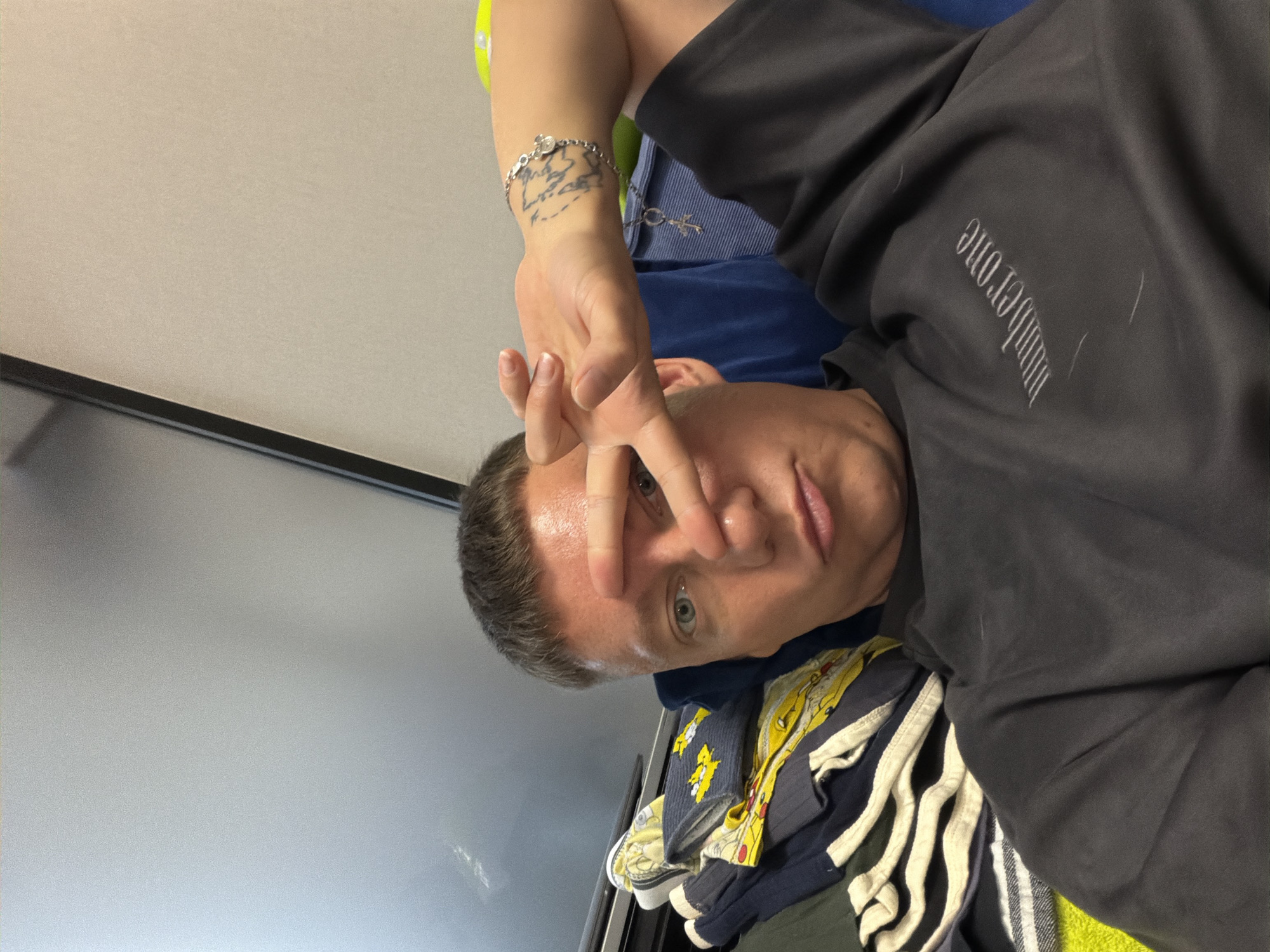 A young man with short brown hair and blue eyes is taking a selfie. He is wearing a dark t-shirt with the words "number one" embroidered on it. He is holding his right hand up to his face, with his index and middle fingers extended in a peace sign. He is also wearing a bracelet with a charm on it. In the background, there is a pile of clothes and a blue cushion. The setting appears to be indoors. There is no visual information to determine the location within the city of Moscow, Russia.