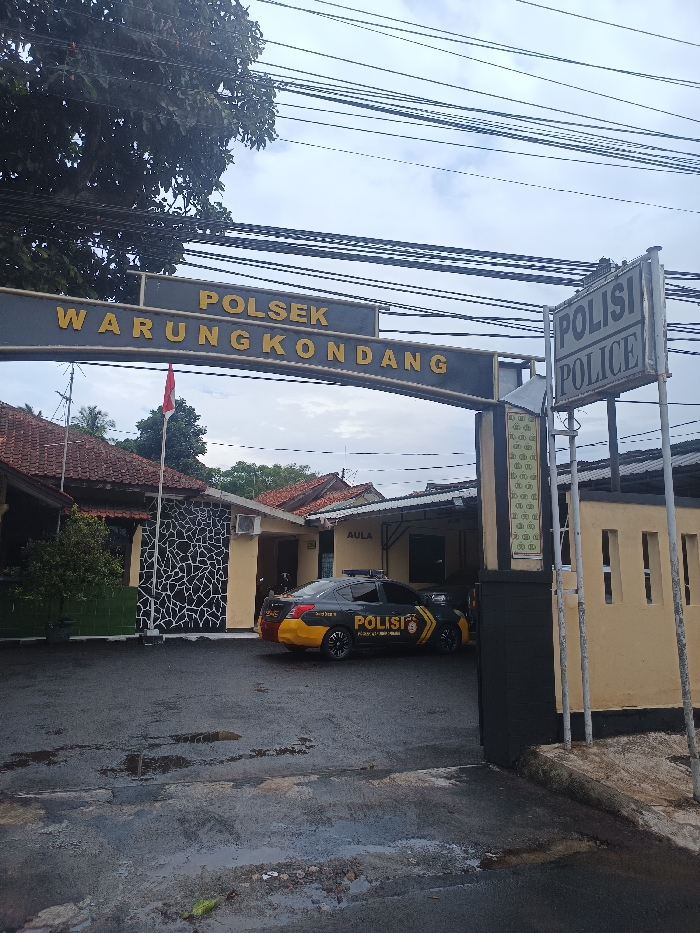 This image displays the exterior entrance of a police station under an overcast sky. A large, dark archway sign, central to the upper half of the frame, bears the gold capital lettering "POLSEK WARUNGKONDANG." To the right of the archway, a vertical pole sign reads "POLISI" above "POLICE" in English. Behind the archway and to the left, an Indonesian flag (red and white) is visible on a flagpole. The station complex consists of several single-story buildings with varying rooflines. A central building has a light yellow exterior with an air conditioning unit and a sign reading "AULA" above an entrance. Parked in front of this building is a black and yellow Indonesian police sedan, equipped with blue roof lights and marked with "POLISI" in white lettering on its side. To the left, another building features a wall section with a black and white abstract pattern. The ground in the foreground is dark, wet asphalt or concrete with visible puddles. Numerous black utility wires are stretched across the sky above the archway. No individuals are present in the scene, and no specific actions or interactions are occurring. The location, as identified by the prominent signage, is Polsek Warungkondang, which is situated in the district of Warungkondang, Cianjur Regency, West Java, Indonesia, not Cikaroya.