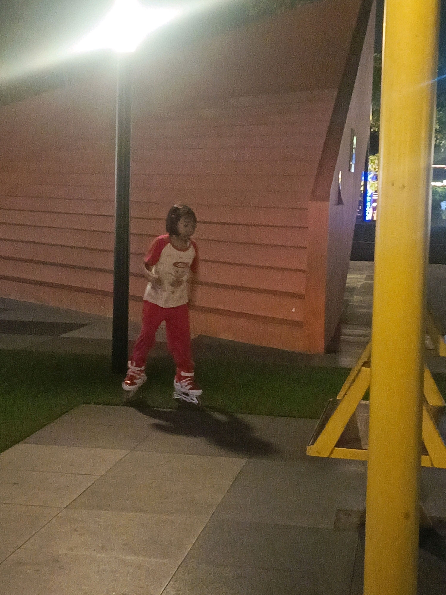 An outdoor shot captures a young child on roller skates in a paved area next to a grassy patch. The child, wearing red pants and a t-shirt with red sleeves, is in motion, with their body angled towards the right of the frame. The background features a textured, horizontally-lined salmon-colored wall and a bright yellow pole. A lamppost with an illuminated light is visible to the left of the child. The scene appears to be at dusk or at night, given the artificial lighting. The image may be from a public area or a park in Sidoarjo, Indonesia.