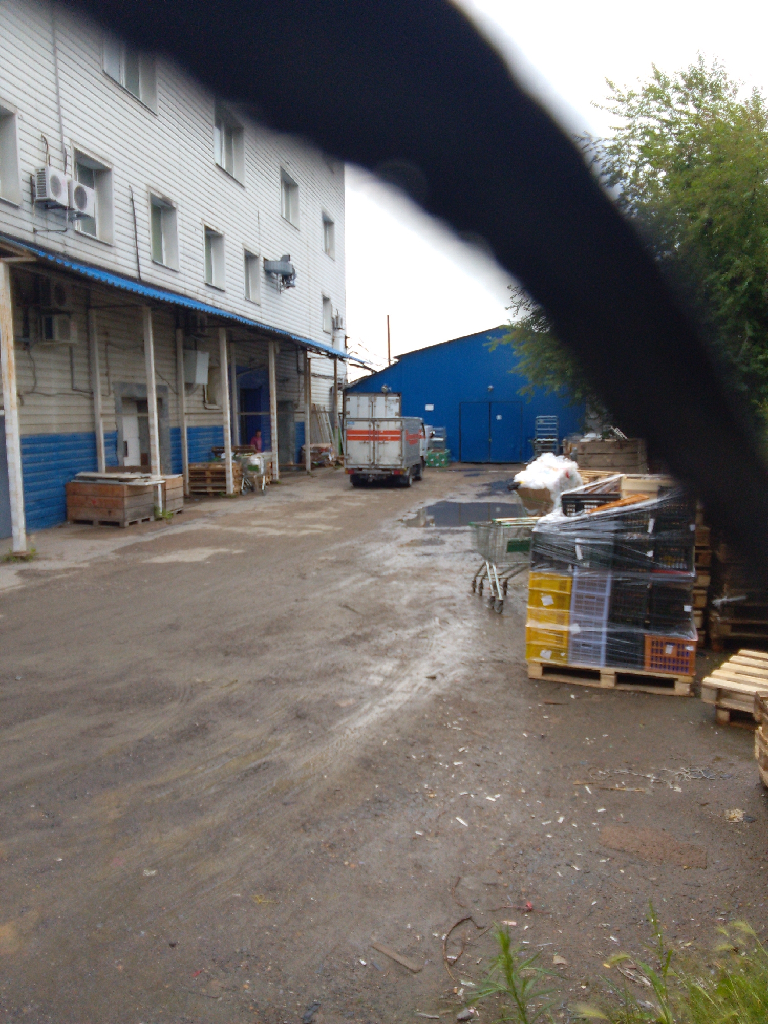 The image depicts an outdoor industrial or storage area, possibly near a logistics facility, in Khabarovsk, Russia. A two-story building with white siding and blue trim is visible on the left, featuring multiple windows and air conditioning units. A covered walkway with blue roofing runs along the base of the building. In front of the building, there are wooden crates and pallets. Further into the scene, a white truck with red stripes is parked on a wet, gravelly surface. Adjacent to the truck, a large blue building with double doors is visible. To the right of the truck, a pile of white material is present, along with stacks of plastic crates wrapped in plastic sheeting, some of which are yellow, grey, and black. Wooden pallets are also scattered throughout the area. A shopping cart is positioned near the stacks of crates. The ground appears wet and somewhat muddy, suggesting recent rain. Trees are visible in the background, with foliage indicating a temperate climate. The overall scene suggests an operational area for storage and transport.