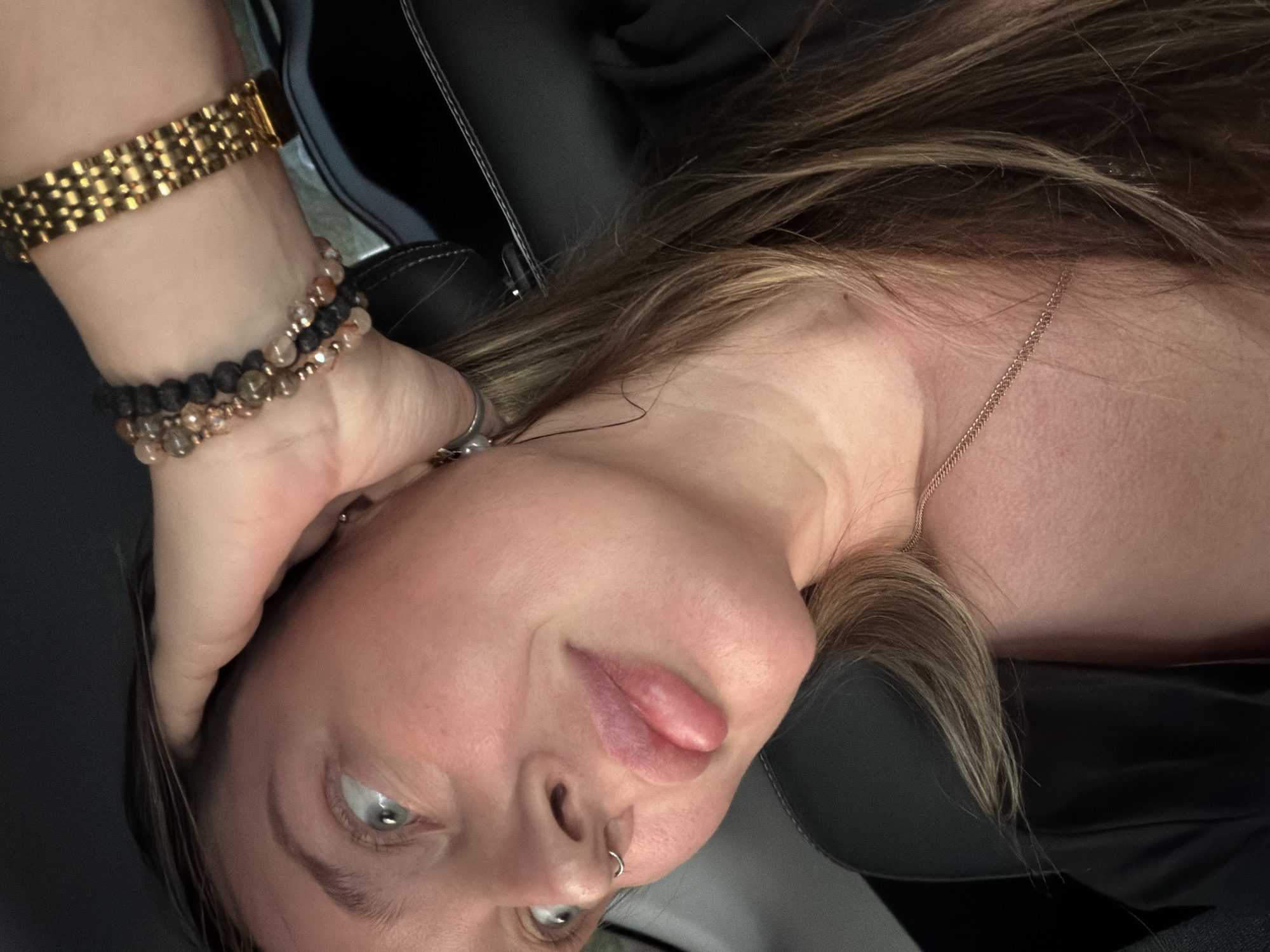 A close-up, high-angle portrait captures an individual, likely female, seated within a vehicle. The subject's left facial profile (from the viewer's perspective) is prominently featured, displaying a light-colored eye gazing upwards and to the right. The lips are full, light pink, and slightly parted. A silver-colored septum ring pierces the nose. The right hand is positioned on the upper left side of the head, with fingers gently contacting brown hair that exhibits lighter streaks. The right wrist is adorned with a wide, gold-colored metal bracelet featuring a grid-like pattern, accompanied by multiple beaded bracelets consisting of dark and translucent stones with metallic accents. A thin gold-colored chain necklace is visible around the neck. The subject wears a dark-colored garment. The background consists of a dark vehicle interior, including a dark seat with visible stitching and a black seatbelt with white stitching crossing the shoulder. The illumination suggests ambient light from the vehicle's exterior. There are no visual elements within this image that provide information or cues pertaining to the specific geographic location of Domodedovo, Russia.