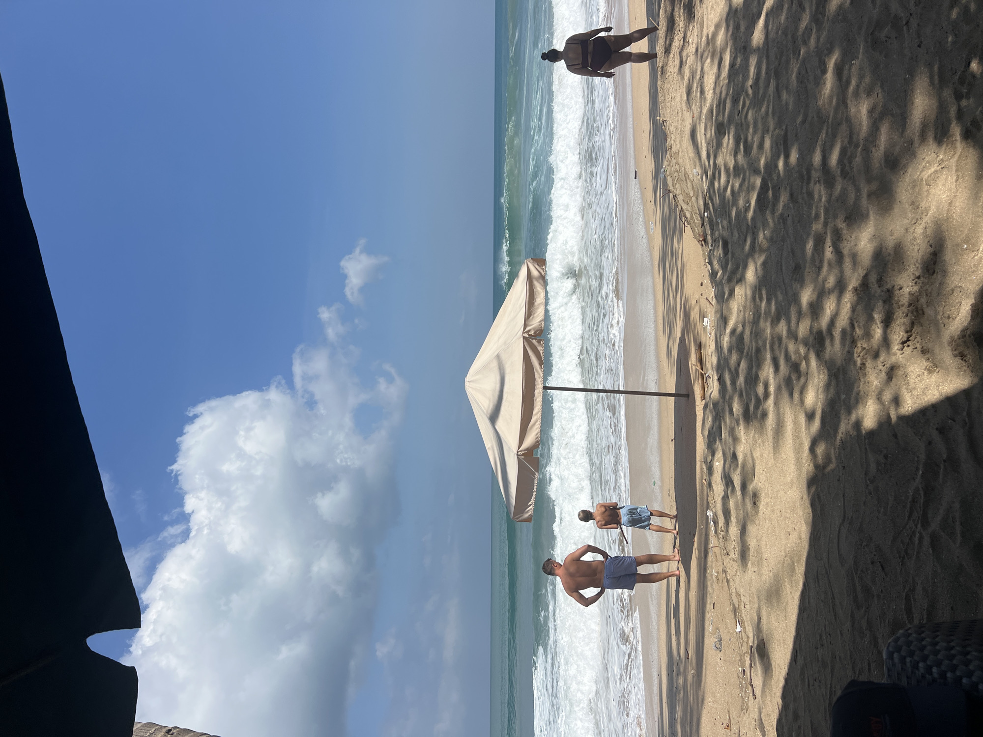 In Kuta, Indonesia, on a sandy beach, two individuals, a man and a young boy, stand facing the ocean. The man, wearing blue checkered shorts, has his back to the camera with his hands on his hips. The boy, in blue patterned shorts, stands to his right, holding a stick. A beige beach umbrella stands between them and the water. Further to the right, a woman in a dark swimsuit faces the ocean, her back to the camera. The sea has moderate waves, and the sky is blue with scattered white clouds. The foreground is a sandy beach with shadows cast from overhead foliage.