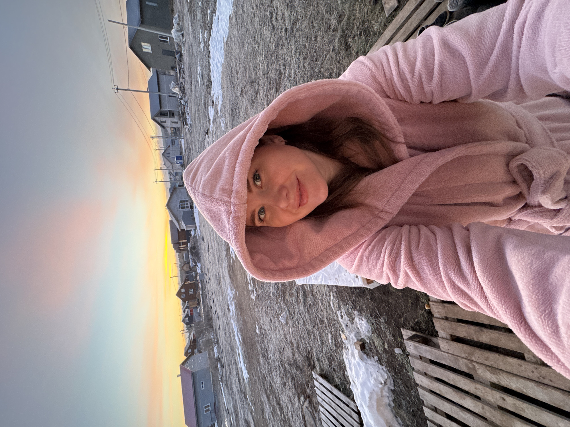 A woman in a pink hooded robe is facing the camera. The setting is Nestiukovo, Russia, recognizable by the style of buildings in the background and known geographic characteristics. Snow patches are present on the ground, suggesting a cold climate. A wooden pallet is located on the right. The sky shows a sunset with a gradient of colors, ranging from yellow to grey.
