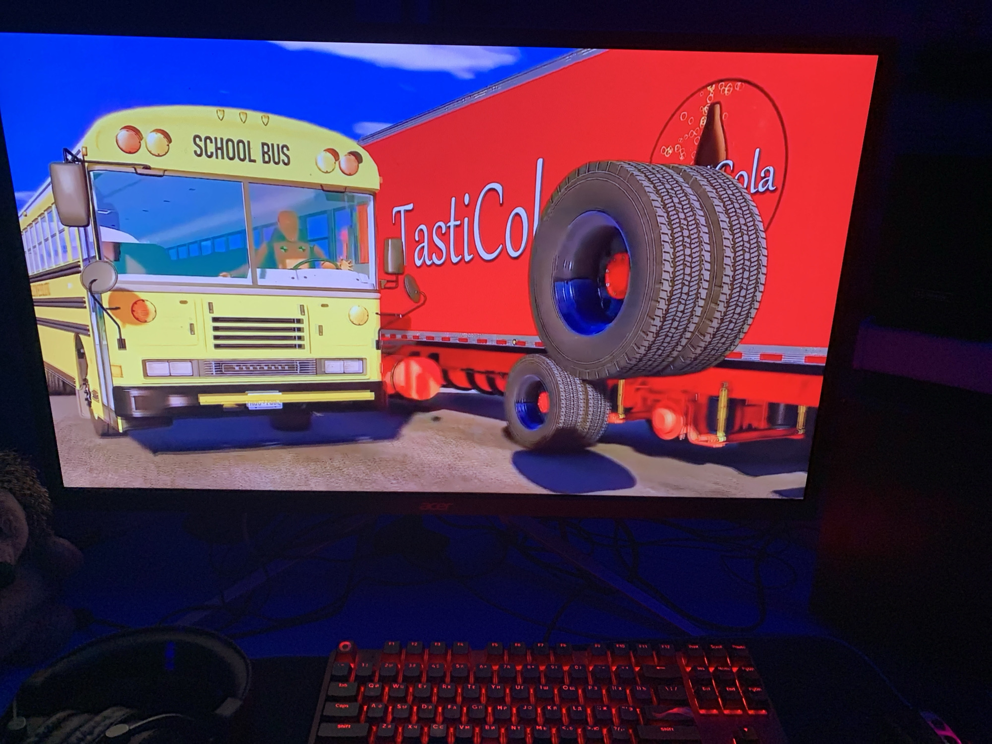 The image shows a computer monitor displaying a scene from what appears to be an animated film or video game. The scene features a yellow school bus next to a red truck with the words "TastiCola" and a logo resembling Coca-Cola on its side. A cartoon character, possibly a mouse, is seen driving the bus. The monitor is positioned on a desk with a red-lit keyboard and a pair of headphones. The cables connected to the monitor are visible. A plush object is partially visible to the left of the monitor. The Acer logo is visible on the monitor's frame. There is no clear indication of the image location being Severodvinsk, Russia.