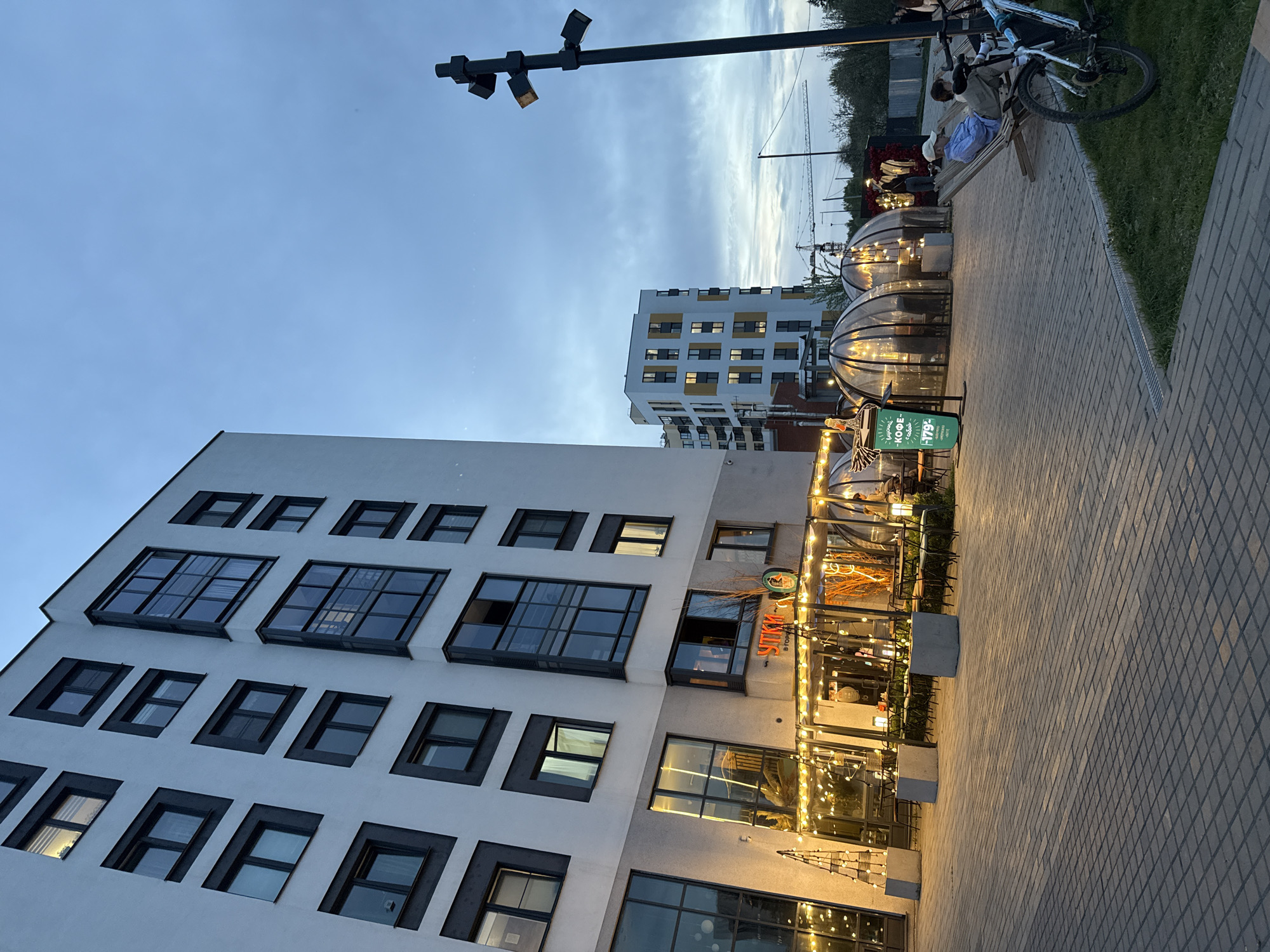 The image depicts an urban scene in Izhevsk, Russia, taken at dusk or twilight. Dominating the foreground is a modern, multi-story building with a white facade and dark-framed windows. Adjacent to the building is a restaurant or cafe, partially enclosed with transparent dome structures illuminated by string lights. Patrons can be seen inside. Outside, a menu board displays a "КОФЕ" (Coffee) offer.
Further along the sidewalk, people are seated on a bench. A bicycle is parked nearby. In the background, a similar, but taller, white building is visible. A construction crane can be spotted in the distance, indicating ongoing development. A street lamp stands prominently at the top left of the frame, adding to the urban landscape. The sky is a blend of light and dark blues, characteristic of twilight. The paving of the walkway is patterned and stretches into the perspective of the shot.