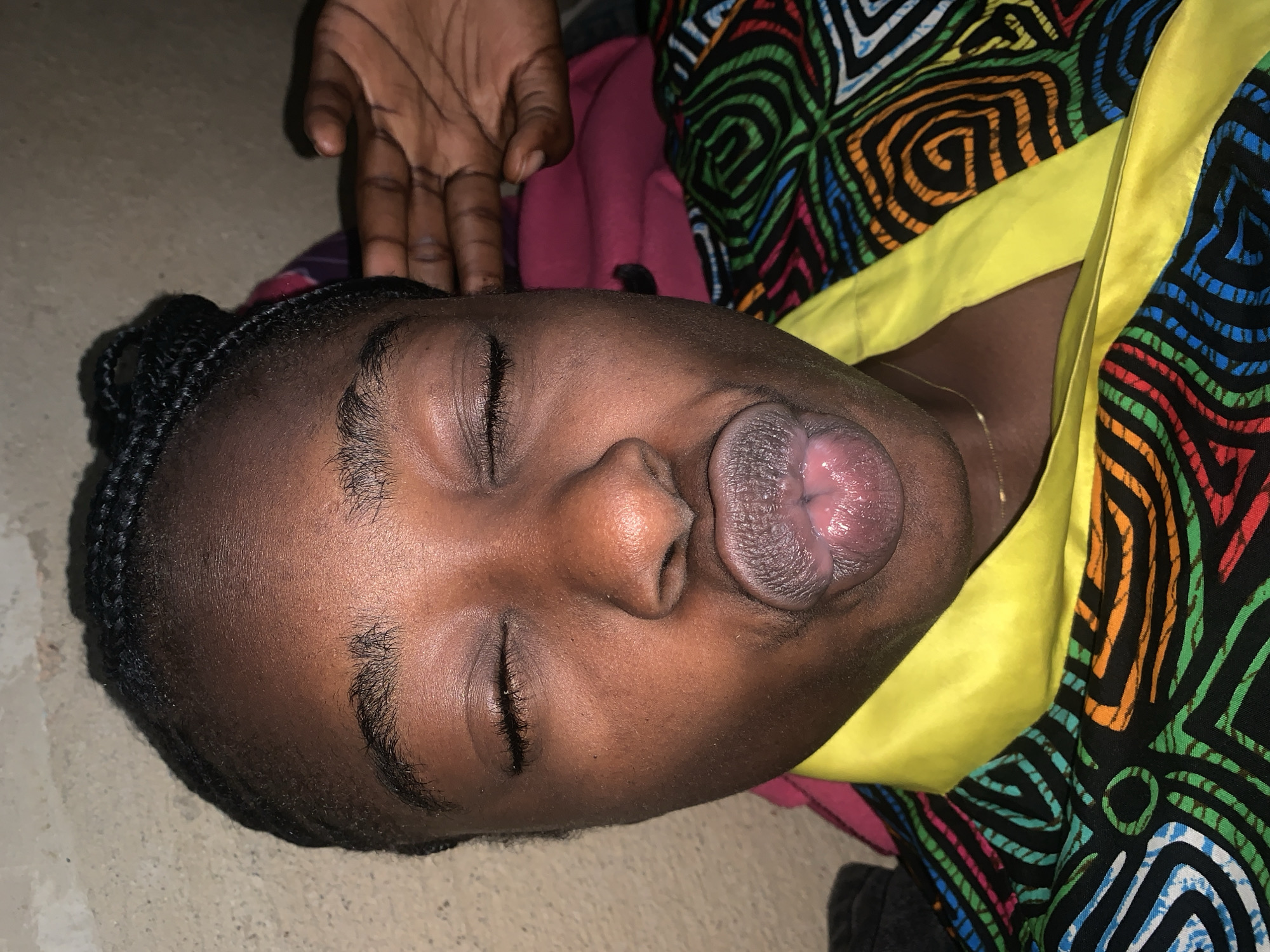 The image is a close-up, high-angle selfie of a young Black woman with her eyes closed and lips pursed. She has braided hair. She is wearing a brightly colored patterned garment with a yellow collar. A hand is visible on the right side of the frame, touching her ear. The background appears to be a textured wall. The scene is indoors, in what could be a residential setting. The specific location in Rafin Par, Nigeria, cannot be determined from the visual information provided.
