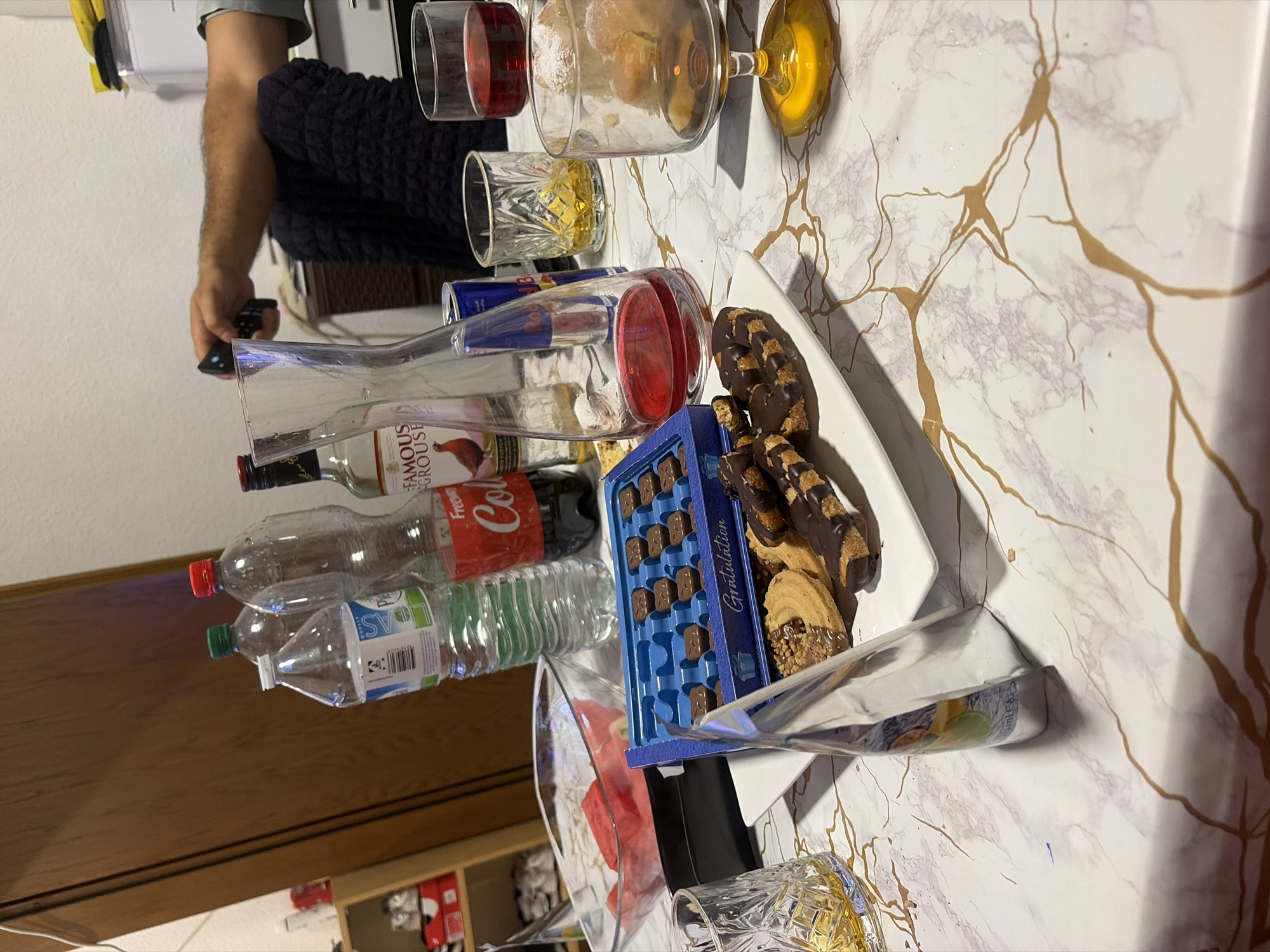 The image depicts a table covered in a marble-patterned tablecloth, laden with various items suggesting a gathering or a snack. Positioned on the table are several bottles of beverages, including a bottle of Coca-Cola, a bottle of Famous Grouse Scotch whisky, and multiple water bottles. There is also a can of Red Bull. A glass decanter filled with a red liquid, likely wine, is present, alongside a wine glass also containing red wine. Another glass contains a golden liquid, possibly whiskey or liquor. A bowl of sliced watermelon sits on a black object, possibly a tray or mat. In front of the drinks and watermelon, a blue box labeled "Gratulation" contains an assortment of chocolates. Adjacent to the chocolate box are several cookies, some topped with chocolate and nuts. A glass with a patterned design is on the left side of the frame, containing a golden liquid. A silver-wrapped item, possibly a food product or condiment, is propped up on its side. In the background, a person's arm and hand are visible, holding a black remote control, suggesting they may be interacting with an electronic device. The environment appears to be indoors, with a wooden cabinet or door visible on the left. The presence of various food and drink items, as well as a decorative box and cookies, indicates a casual setting. The specific location in the city of Duisburg, Germany, cannot be determined from the visual information provided.
