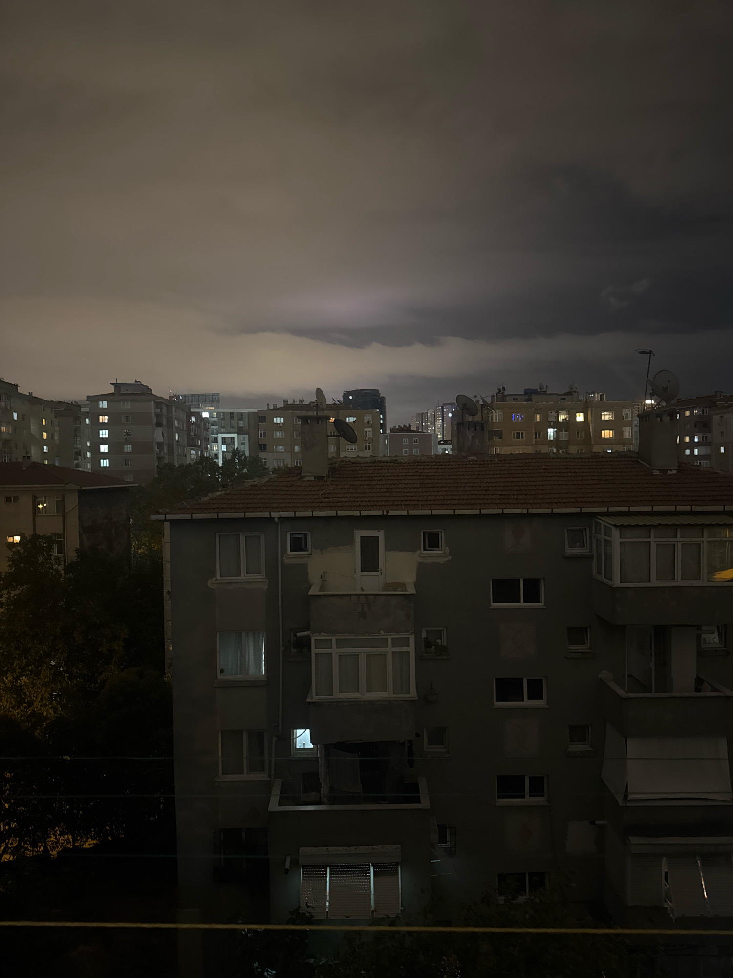The image depicts a nighttime urban scene in Gungoren, Turkey, characterized by a dramatic, cloudy sky. The foreground features the facade of a weathered apartment building, with several windows visible, some illuminated from within, casting a soft glow into the dark. The building's architecture suggests a utilitarian, older style, with a prominent tiled roof and a protruding balcony on the right side.
Beyond this building, the cityscape stretches into the distance, with multiple apartment blocks of varying heights and designs. Many of these buildings also have illuminated windows, suggesting residential activity. The sky above is filled with heavy, dark clouds, hinting at stormy weather. Faint lightning can be observed in the far distance, illuminating the clouds from within, adding a sense of atmospheric tension to the scene.
The overall mood is somber and slightly ominous due to the dark sky and the limited visibility. The presence of trees on the left side of the image provides a touch of natural element amidst the urban sprawl. No people are discernible in the image. The time of day is clearly night, and the weather appears to be a storm approaching or passing. The location is indicated as Gungoren, Turkey, which aligns with the visual cues of dense residential apartment buildings common in many Turkish cities. No visible text is present in the image.