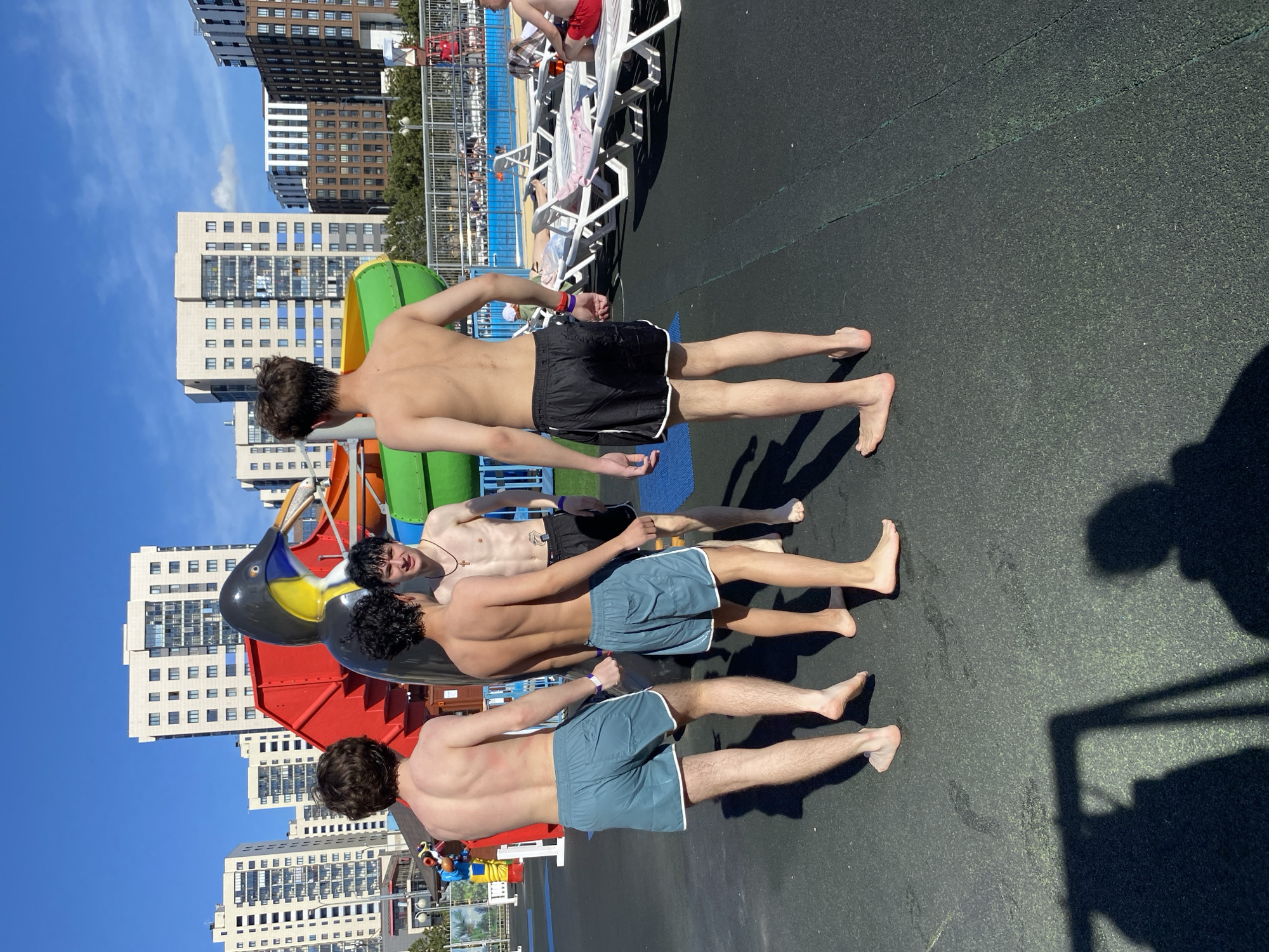 Four shirtless young men stand in a line outdoors, possibly at a water park. The first man wears black swim trunks, while the other two wear light blue ones. The fourth man’s swimwear is not visible. Behind them, there is a colorful water slide structure. In the background are high-rise buildings, suggesting an urban location. Other people are visible in the background sunbathing on lounge chairs. The setting appears to be Kazan, Russia, as the architecture and overall environment are consistent with the city.