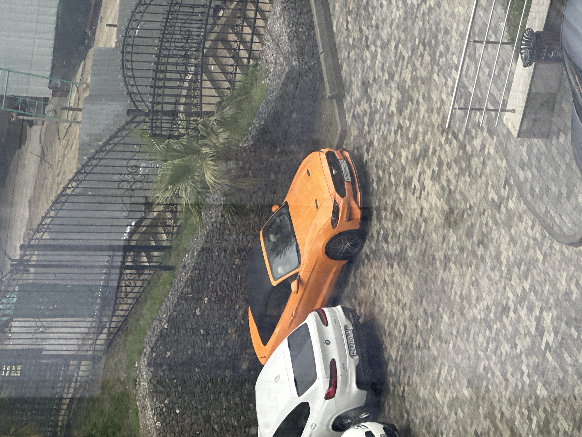 The image shows an aerial view of an outdoor scene in what appears to be the city of Sochi, Russia, based on the license plate. It's a view seen through a window. Two parked cars are visible: a bright orange sports car and a white SUV or crossover vehicle. The orange car is positioned partially on the gravel next to a low retaining wall built with large stones. There's a black wrought-iron gate behind the orange car. The white car is on the paved area. A section of a railing is visible on the top right. The backdrop includes greenery and rooftops, indicating an urban or suburban area. The pavement is made of interlocking gray and white stones.