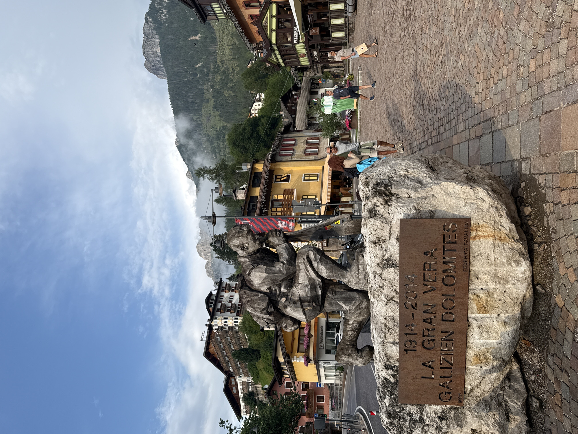 The image depicts a public square in Moena, Italy, featuring a large wooden sculpture of a kneeling soldier. The sculpture is situated on a large rock with a metal plaque that reads "1914-2014 LA GRAN VERA GALIEN DOLOMITES FEDERICA CAVALLIN MSE". In the background, there are buildings characteristic of a European mountain town, including hotels and shops. A prominent hotel with "HOTEL" signage is visible on a hillside. The scene is set against a backdrop of lush green mountains partially obscured by clouds. Several people are walking through the square, dressed in casual attire. The square itself is paved with cobblestones. The sky is blue with scattered clouds.