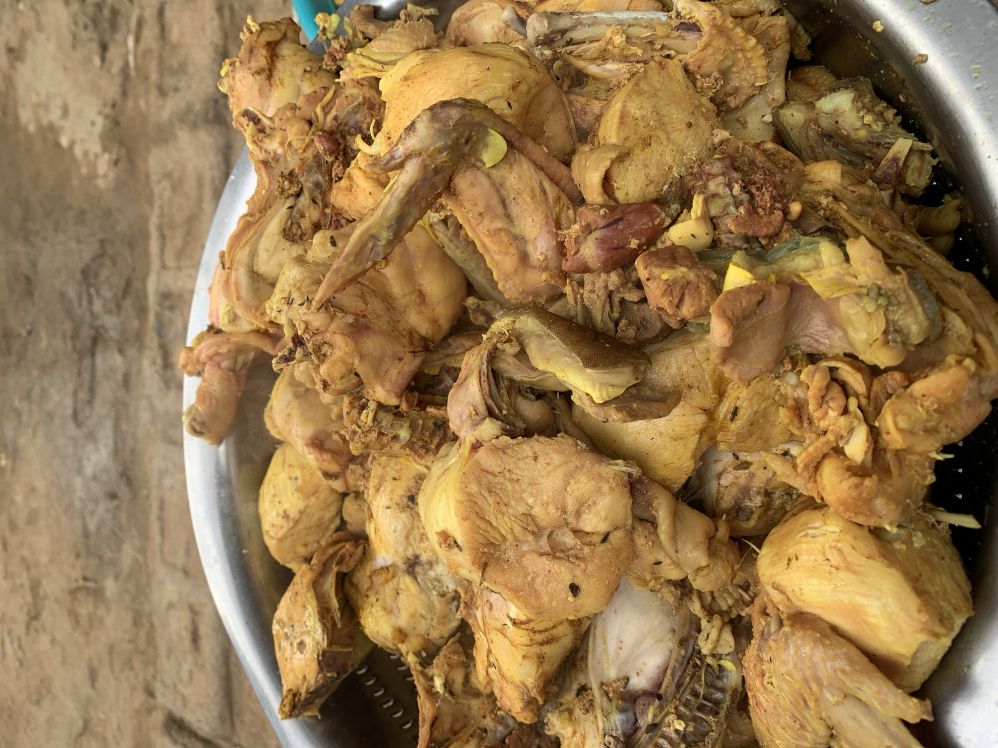 The image displays a high-angle, close-up perspective of a large quantity of chicken pieces contained within a metallic basin. The chicken appears par-cooked or boiled, exhibiting a predominant yellowish-brown color, indicative of seasoning. Small, dark particulate matter, consistent with herbs or ground spices, is visible on the surface of multiple pieces. Identifiable parts include bone-in chicken wings, breast sections, and various other cut fragments, along with visible skin and some organ meat (e.g., a reddish piece resembling a heart). Faint vapor or steam is discernible rising from the chicken, suggesting it is hot. The container is a large, round metallic basin or bowl, with perforations visible along its upper left rim, suggesting it might be a colander or steamer component. The background is out of focus, showing an uneven, earthy-toned surface, possibly dirt or rough concrete, indicating an outdoor or semi-outdoor setting. No individuals, specific actions, or interactions are visible. The scene is contextually located in New Stadium, Nigeria.