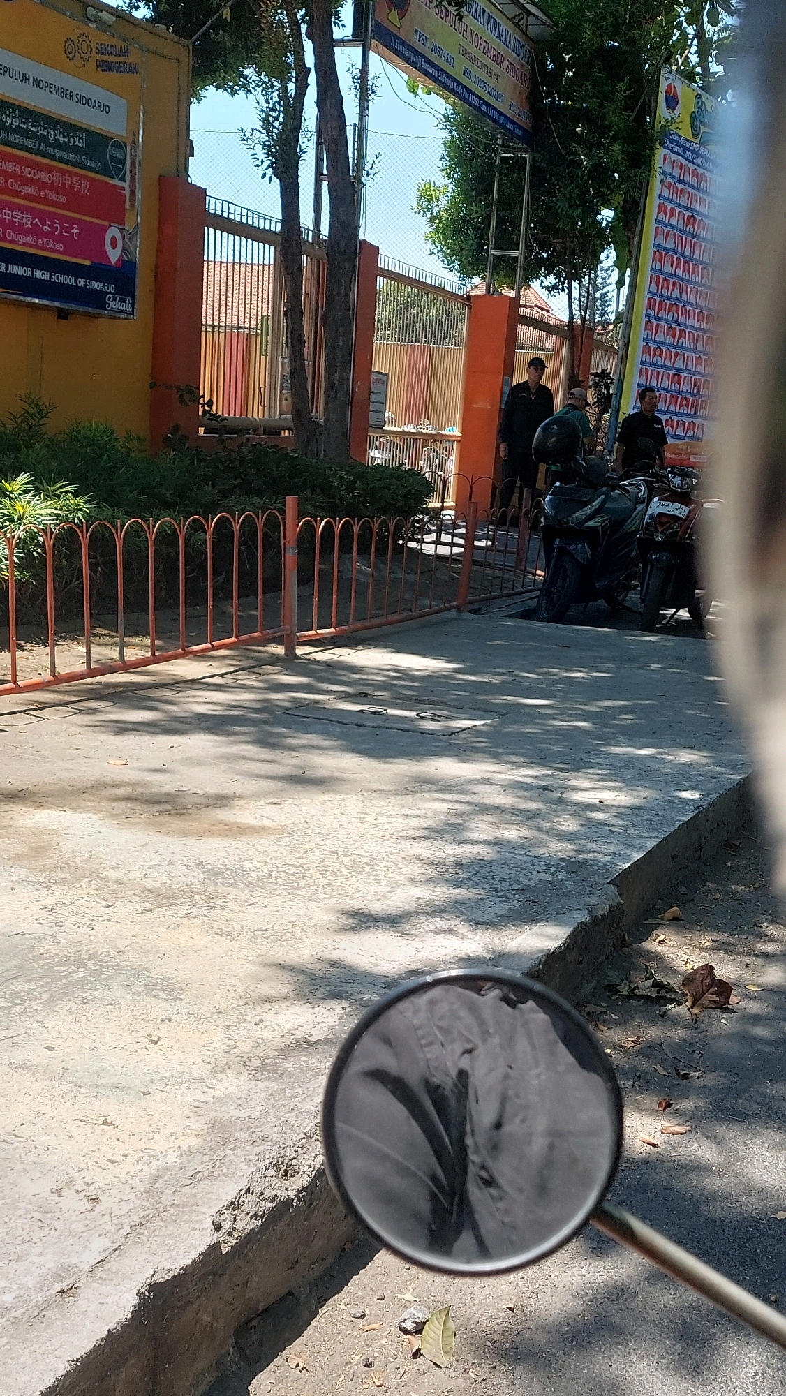The image shows the entrance to "SEKOLAH PEMBEGARAN SEPULUH NOPEMBER SIDOARJO" in Siwalan Panji, Indonesia. A yellow building with an orange gate is visible on the left, displaying signs in various languages, including Arabic, Japanese, and Indonesian. The main sign above the entrance reads "SEKOLAH PEMBEGARAN SEPULUH NOPEMBER SIDOARJO". Further to the right, a taller banner displays rows of small pictures, possibly of students or staff. Three men are standing near the gate, and two motorcycles are parked in front of it. The foreground is a concrete sidewalk with shadows from trees, and a motorcycle's rearview mirror is visible in the bottom right corner, reflecting the scene. The environment appears to be outdoors on a sunny day.