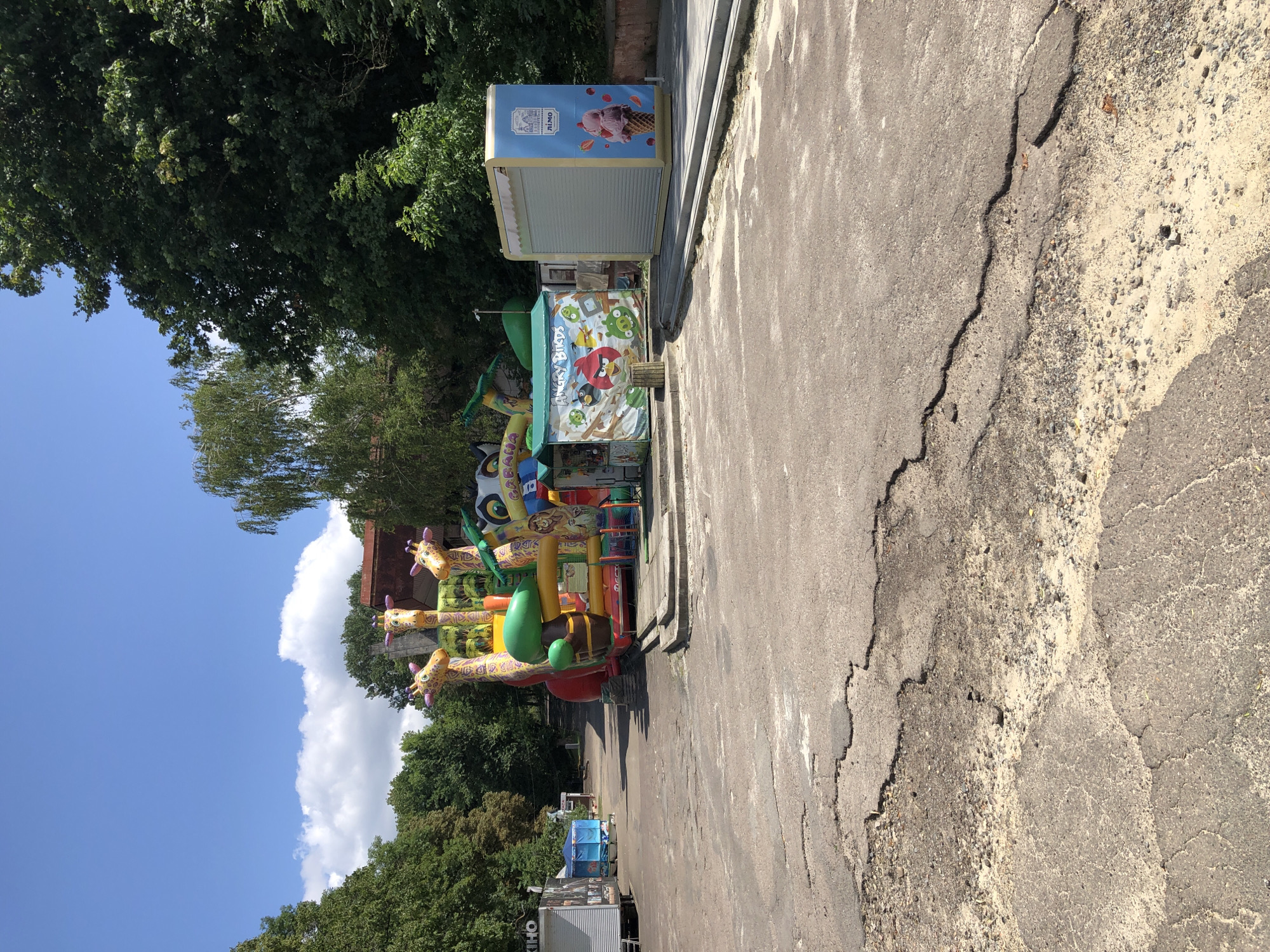 This is an outdoor, eye-level, medium-shot photograph taken on a sunny day in Lviv, Ukraine. The foreground features cracked asphalt and dirt. In the mid-ground, there is a large inflatable playground with giraffe figures, as well as two small kiosks. One kiosk has an "Angry Birds" theme, while the other displays an advertisement for ice cream with the text "LIMO" and Cyrillic script. In the background, there are trees and buildings. The sky is blue with scattered clouds.