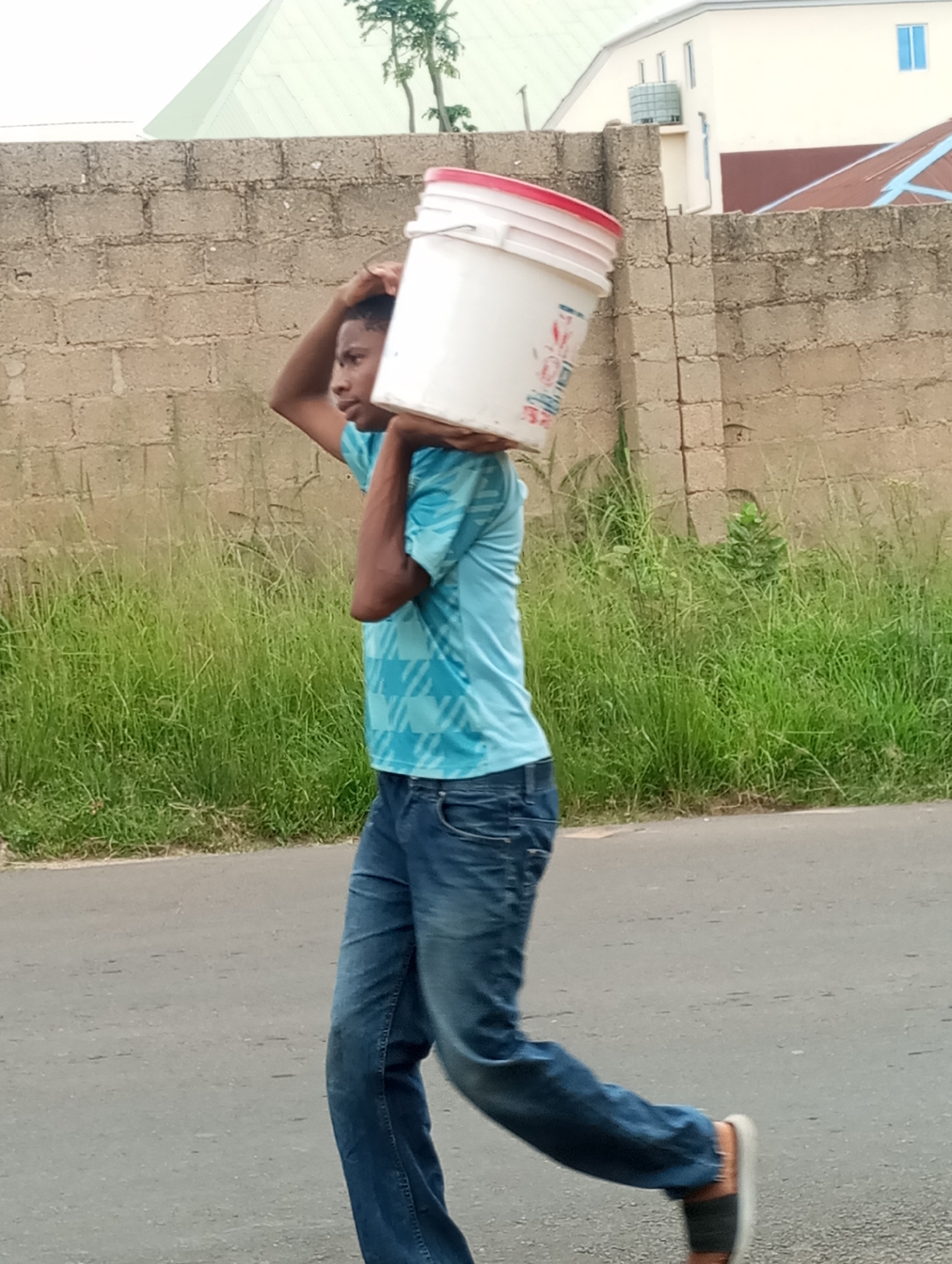 A young man is seen carrying a white bucket with a red rim on his head. He is wearing a light blue t-shirt with a geometric pattern and blue jeans. He is walking on an asphalt road next to a tall, overgrown grassy embankment. Behind the embankment is a brick wall. In the background, there are buildings, including one with a light-colored facade and blue windows, and another with a red roof. The sky is not visible. The scene appears to be in an outdoor urban or semi-urban environment. This image does not provide specific visual cues to pinpoint the exact location within Jos, Nigeria.