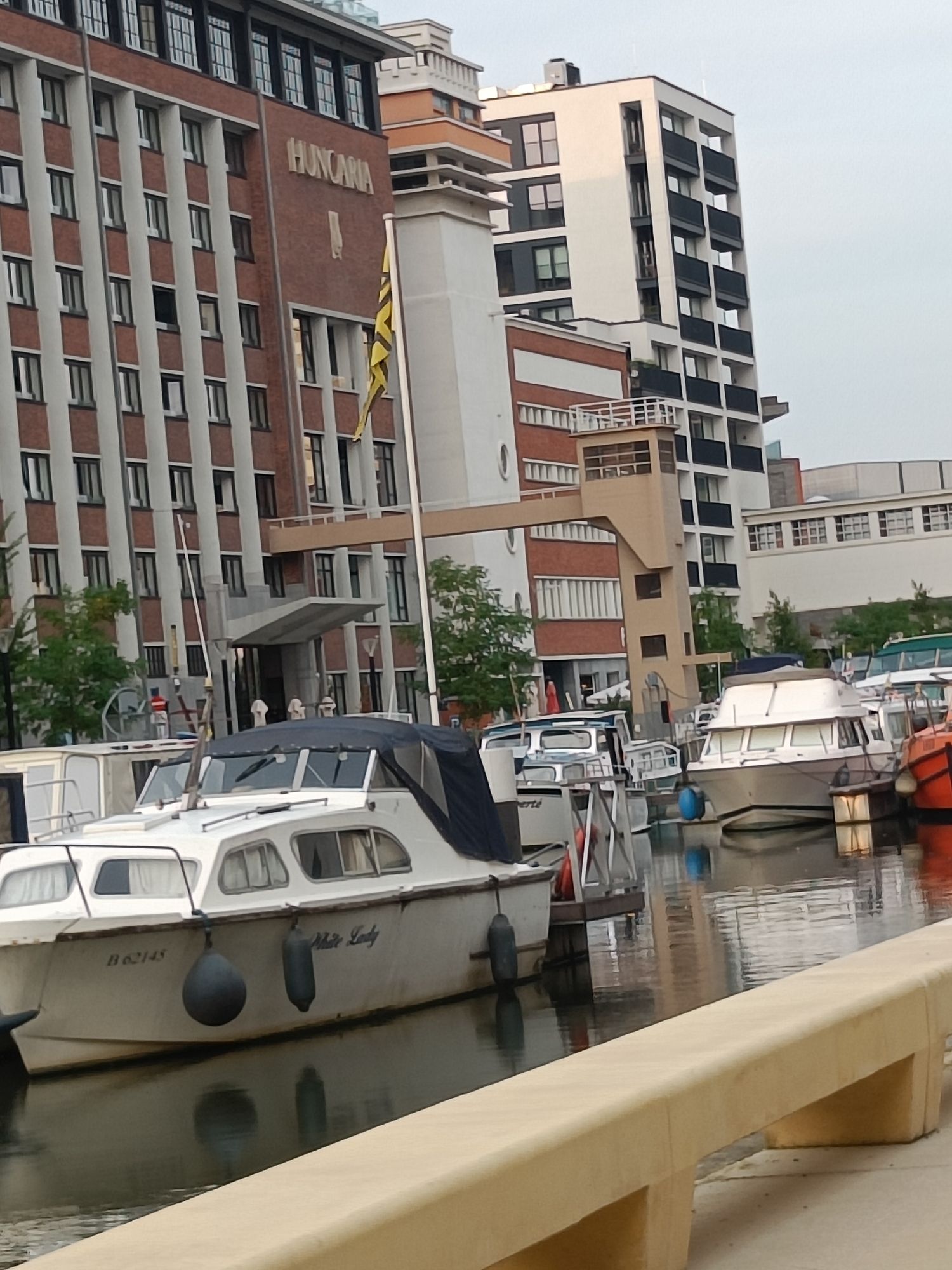 A wide shot shows boats docked in a canal alongside modern buildings. The boats are various sizes and types, with some covered and others open. The buildings are multi-story, with a mix of brick and glass facades, and a distinctive yellow and black striped flag is visible. The water is calm, reflecting the buildings and boats. A light-colored concrete barrier runs along the foreground of the image. The sky is overcast.