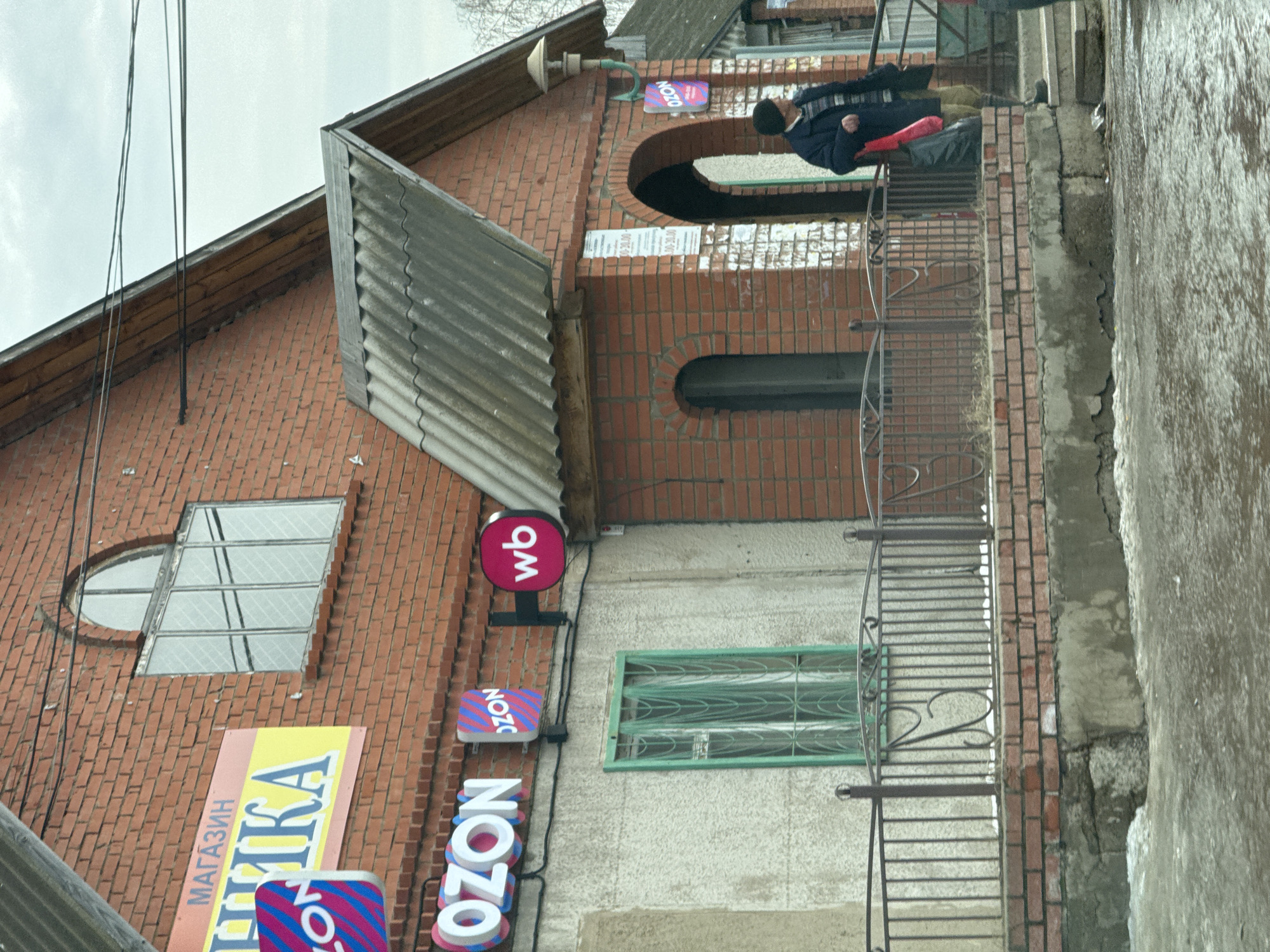 The image depicts a building in Noviy, Russia. The building is primarily constructed of red brick and features a grey metal roof and awning. Signage for "OZON" is visible on the facade, along with a "WB" logo. A person in a dark coat and hat is present on an elevated porch or balcony, holding what appears to be a red bag. The street or sidewalk is wet, and the sky is overcast. Electrical wires are visible in the upper left corner of the image.