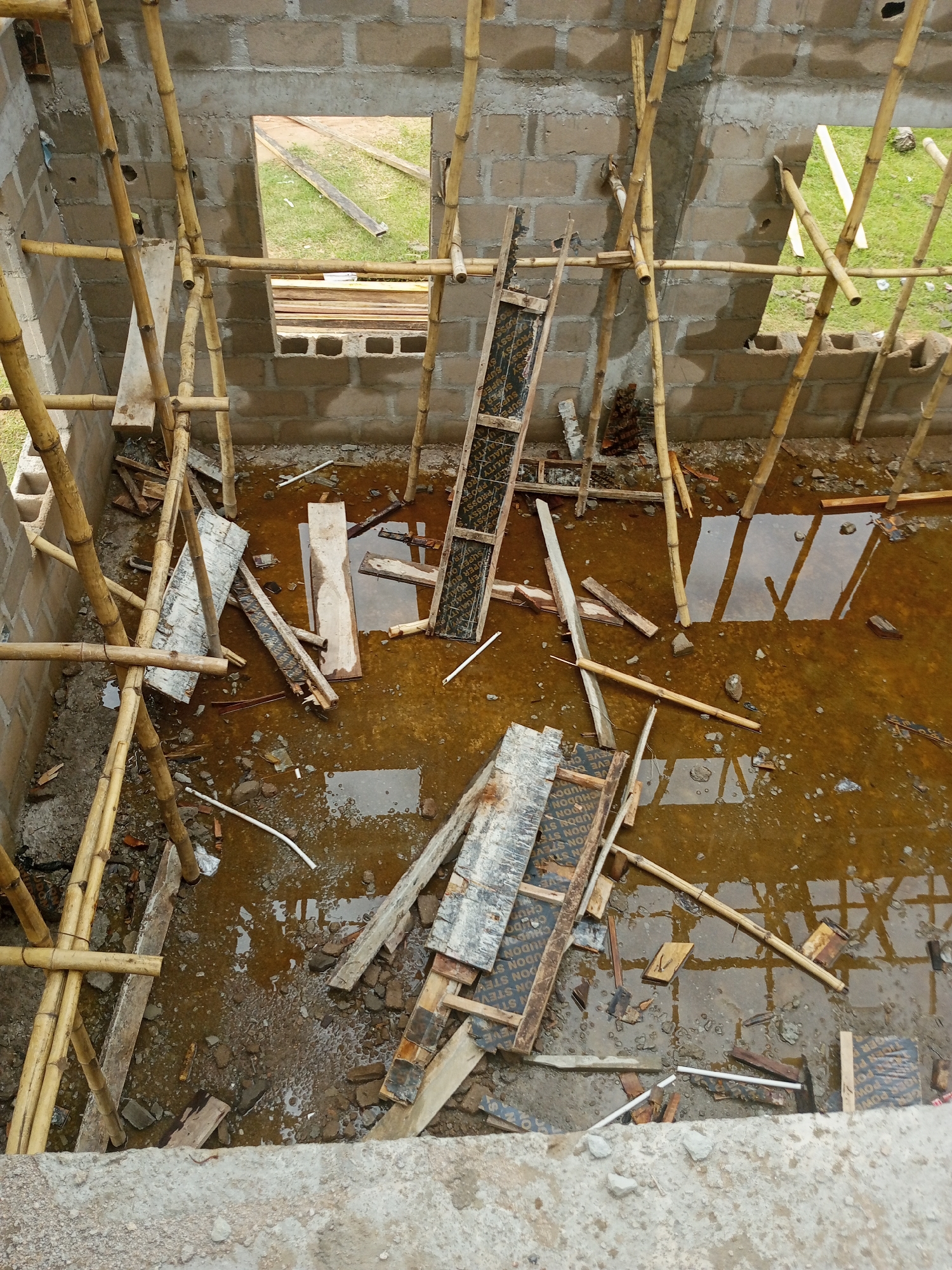 This media file captures an interior view of a building under construction in Jalingo, Nigeria. The scene is characterized by unplastered concrete block walls and a floor covered with muddy water and debris, indicating recent rain or an incomplete drainage system. Scaffolding made of bamboo poles is erected around the structure, supporting wooden planks and construction materials.
Visible objects include various pieces of lumber, plywood sheets, and what appears to be shuttering panels for concrete casting. Some of these panels have text printed on them, with visible phrases like "UGON STEV" and "CHUDON," likely part of branding or instructions. The rough texture of the concrete blocks and the scattered construction waste contribute to the raw, unfinished state of the building.
The presence of standing water and the overall dampness suggest it might be a humid or rainy period in Jalingo. The natural light filtering into the open structure indicates it is daytime, although the exact time is not discernible. No people are visible in the frame, emphasizing the site's current inactive or unattended status. The view is from a slightly elevated perspective, looking down into the construction site.