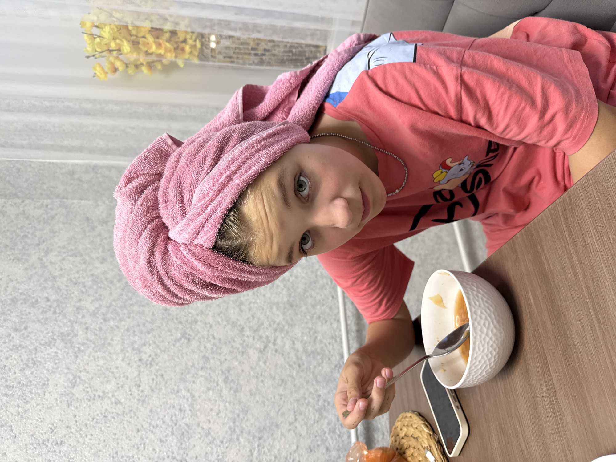 In this close-up, top-down shot, a young girl with a pink towel wrapped around her head is sitting at a wooden table, looking directly at the camera with a curious expression. She is wearing a pink t-shirt with a cartoon cat graphic and a silver necklace. Her right hand is holding a spoon, poised to take food from a white, textured bowl. The bowl contains a light-colored, possibly soupy food. A smartphone and a woven placemat are visible on the table. The background shows a textured gray wall on the left and a blurred glimpse of what appears to be a sofa on the upper right, suggesting an indoor setting, likely a dining area or living room. The lighting appears to be natural, possibly from a window, and the overall mood is casual and domestic. The image is oriented sideways. The girl's expression is neutral, perhaps slightly surprised or amused by the camera. No specific time of day or weather conditions are discernible, but the indoor setting implies a comfortable environment. The location is Stary Oskol, Russia.