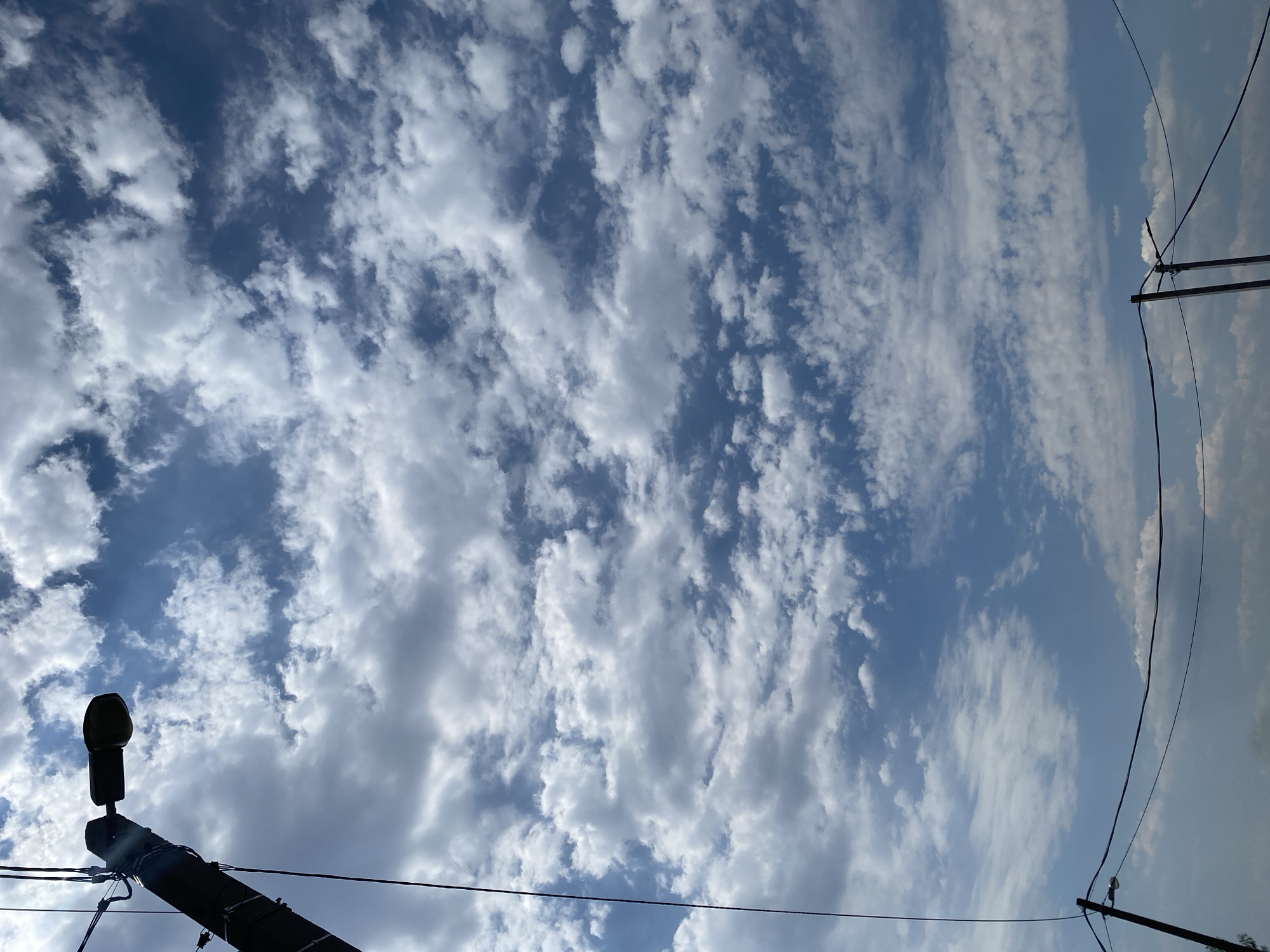 An upward-facing shot shows a bright blue sky filled with scattered white cumulus clouds. A dark silhouette of a utility pole with a street lamp extends from the left edge of the frame upwards, with electrical wires strung across the lower portion of the image. In the distance, another utility pole with a street lamp is visible. The scene appears to be set in an outdoor urban environment, likely in Chornomorsk, Ukraine, given the sky and typical urban infrastructure.