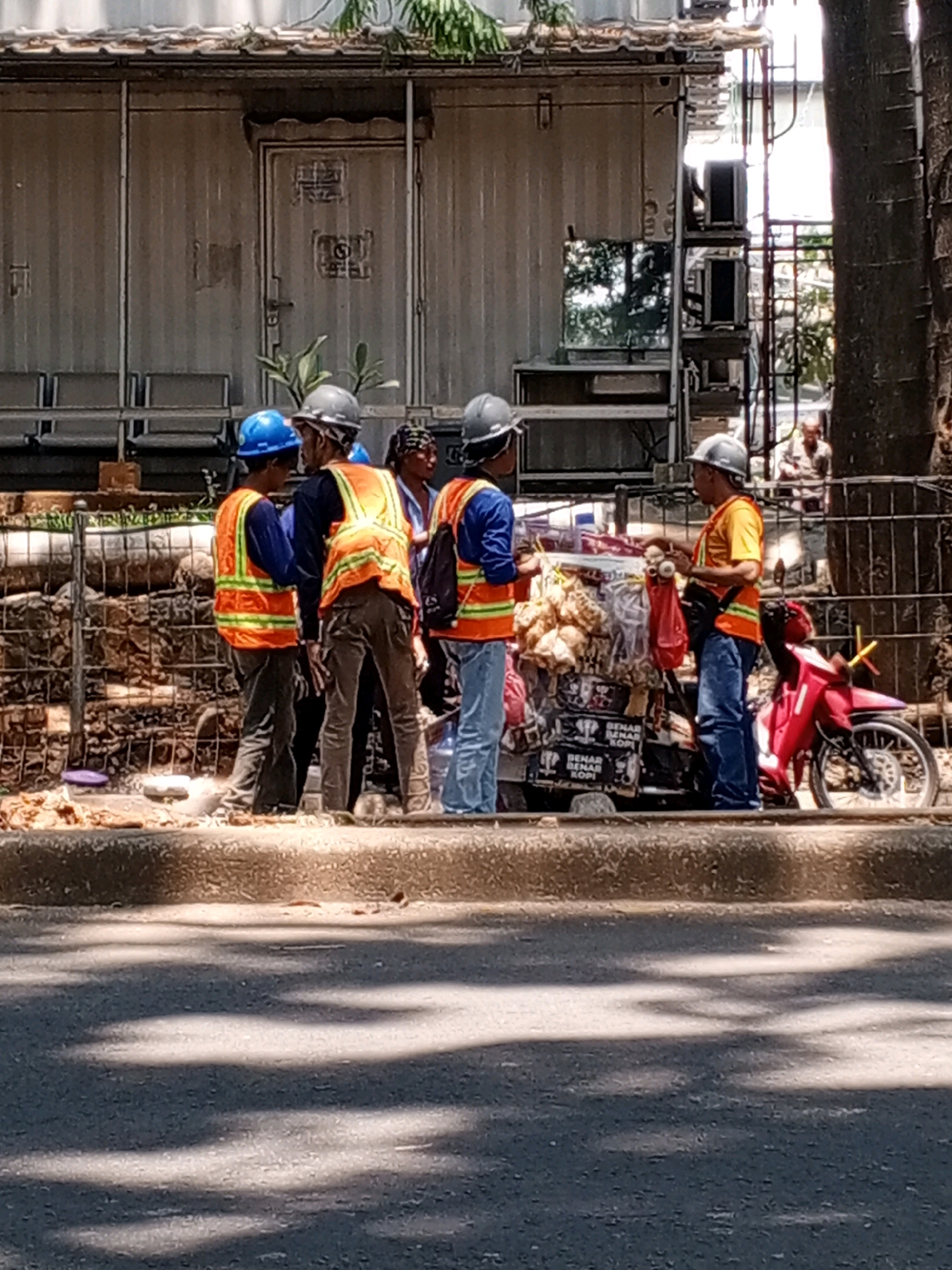 The image depicts a street scene in South Tangerang, Indonesia. In the foreground, a paved road is visible with dappled shadows from trees. A raised concrete curb separates the road from a sidewalk area. On the sidewalk, a group of five individuals in construction attire, wearing high-visibility vests and hard hats, are gathered. One individual, wearing a blue hard hat, stands facing away from the camera, while another wearing a gray hard hat is partially visible in the center. Two individuals in orange high-visibility vests are interacting with a street vendor's cart, which is laden with packaged goods, possibly snacks or drinks. The vendor, wearing an orange vest and blue jeans, is serving them. A red motorcycle is parked to the right of the cart. Behind the group, a corrugated metal structure with a door and windows is visible, likely a temporary building or container at a construction site. In the background, a man is seated further down the street. The overall environment suggests a busy urban area undergoing construction or development.