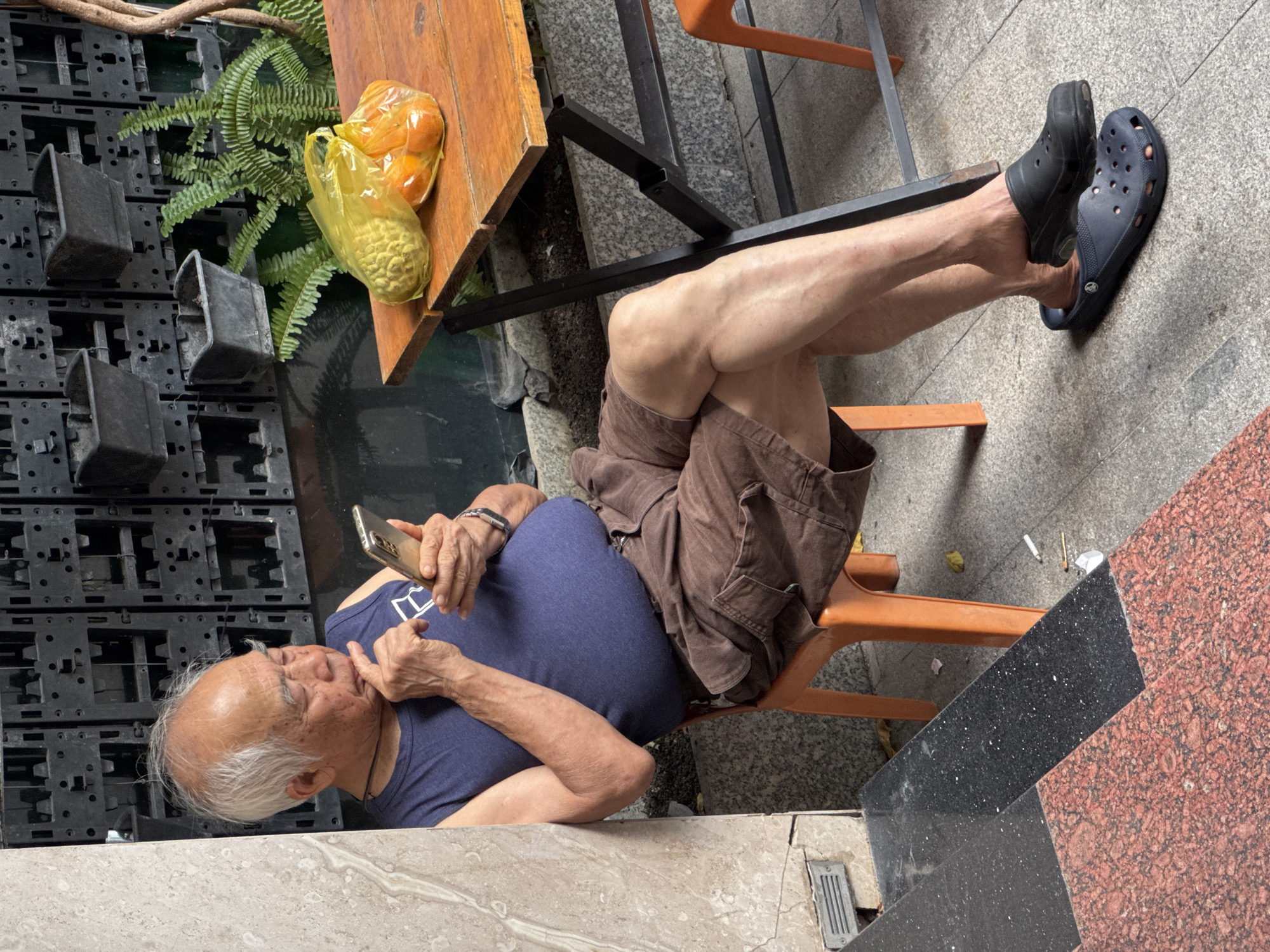 In Nha Trang Ward, Vietnam, an elderly man with white hair and a blue sleeveless shirt is seated on an orange plastic chair, leaning against a light-colored marbled pillar. He is holding a smartphone in his right hand, with his left hand resting on his chin, appearing contemplative. He is wearing brown cargo shorts and dark clogs on his feet. To his right, a wooden table is set up with a yellow plastic bag containing what appear to be fruits, possibly oranges or tangerines. Behind the table, a dark wall with a grid of planter boxes and a fern are visible. The ground is paved with speckled stone tiles, and discarded cigarette butts are present on the ground near the man's feet. The overall scene suggests an outdoor seating area, possibly a cafe or street-side rest spot.