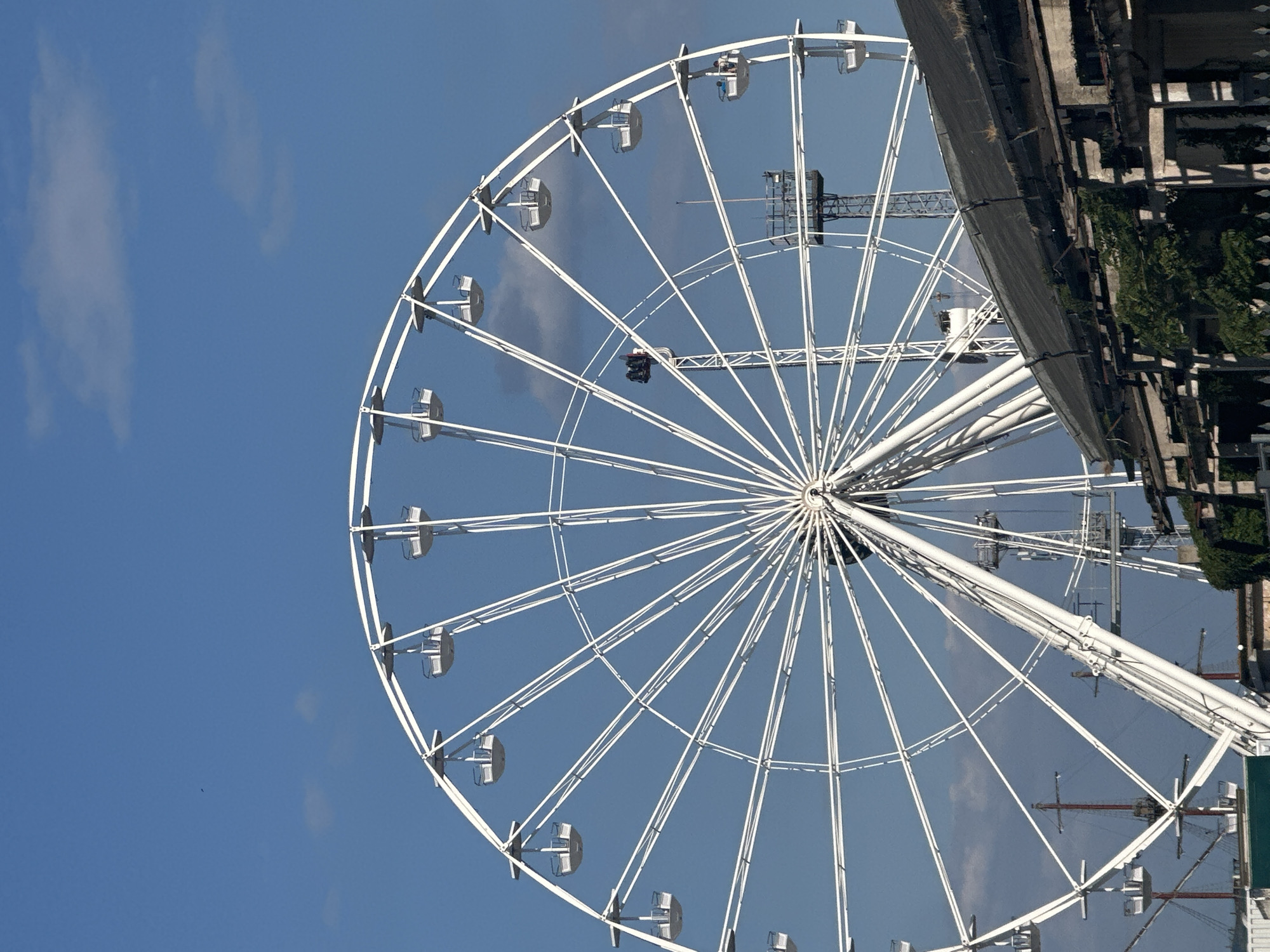 An outdoor, eye-level, vertical, medium shot captures a large, white Ferris wheel against a bright blue sky dotted with scattered clouds. The Ferris wheel dominates the upper two-thirds of the frame. Its intricate white structure is composed of numerous spokes radiating from a central hub, with small, white gondolas suspended from the outer rim. The lower portion of the image features the roofline of a dilapidated building, with dark, weathered shingles and exposed concrete structural elements. Green ivy and other vegetation are creeping up the side of the building. In the distance, behind the Ferris wheel, are two tall, dark, lattice-like towers and what appears to be the masts of a ship. The overall impression is of an amusement park or waterfront area, possibly undergoing renovation or in a state of disrepair.