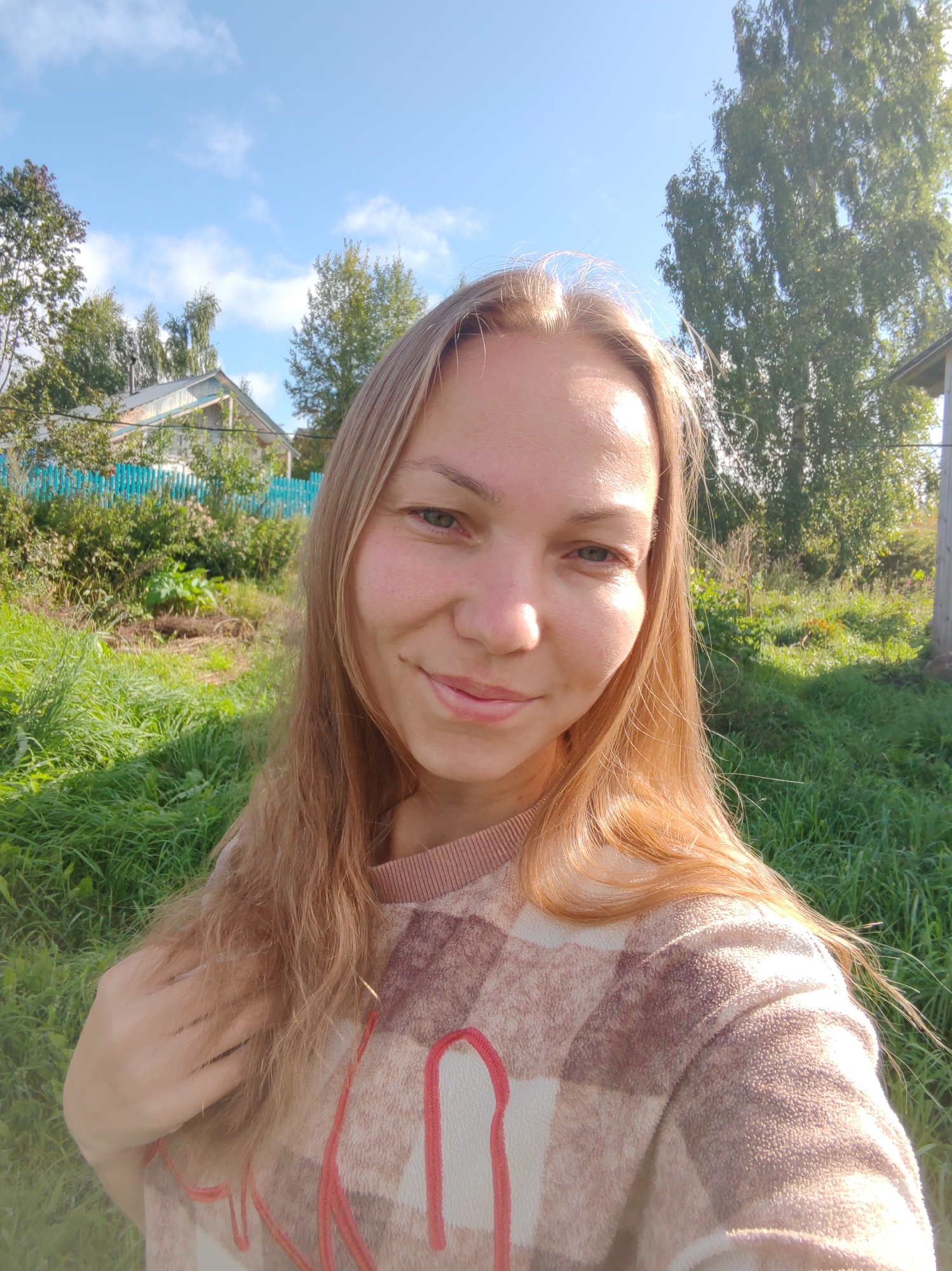 A young woman with blonde hair and green eyes smiles at the camera, her face positioned slightly off-center. She is wearing a long-sleeved, plaid-patterned sweater in shades of brown and white with red accents. The background suggests a rural or semi-rural setting. Lush green grass covers the foreground, with various trees and vegetation behind it. In the middle ground, a light-colored house with a dark roof and a turquoise wooden fence are visible. The sky above is blue with scattered white clouds. The scene appears to be an outdoor photograph taken during daylight, possibly in a garden or yard. The location is identified as Chukhlem, Russia.