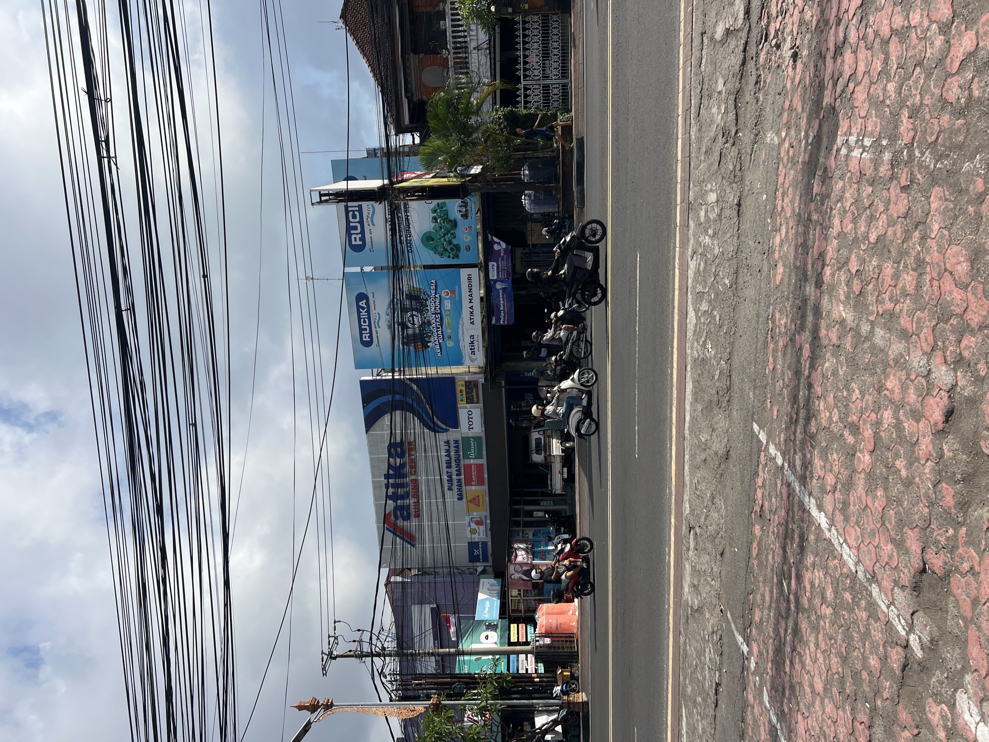 This image captures a street scene in Denpasar, Indonesia, likely on a commercial street with multiple businesses. The foreground shows a portion of a road and a damaged sidewalk with red hexagonal pavers. In the midground, several motorcycles are being ridden on the road. On the right, a man is walking on the sidewalk. Behind the road, a row of buildings is visible. The largest building is "ATIKA BUILDING CENTER", which also has signs for "PUSAT BELANJA BAHAN BANGUNAN" and various product brands like "TOTO". To the right of "ATIKA BUILDING CENTER" are buildings with large blue billboards advertising "RUCKA". The buildings are adorned with numerous overhead electrical wires, typical of urban Asian infrastructure. The sky is partly cloudy.