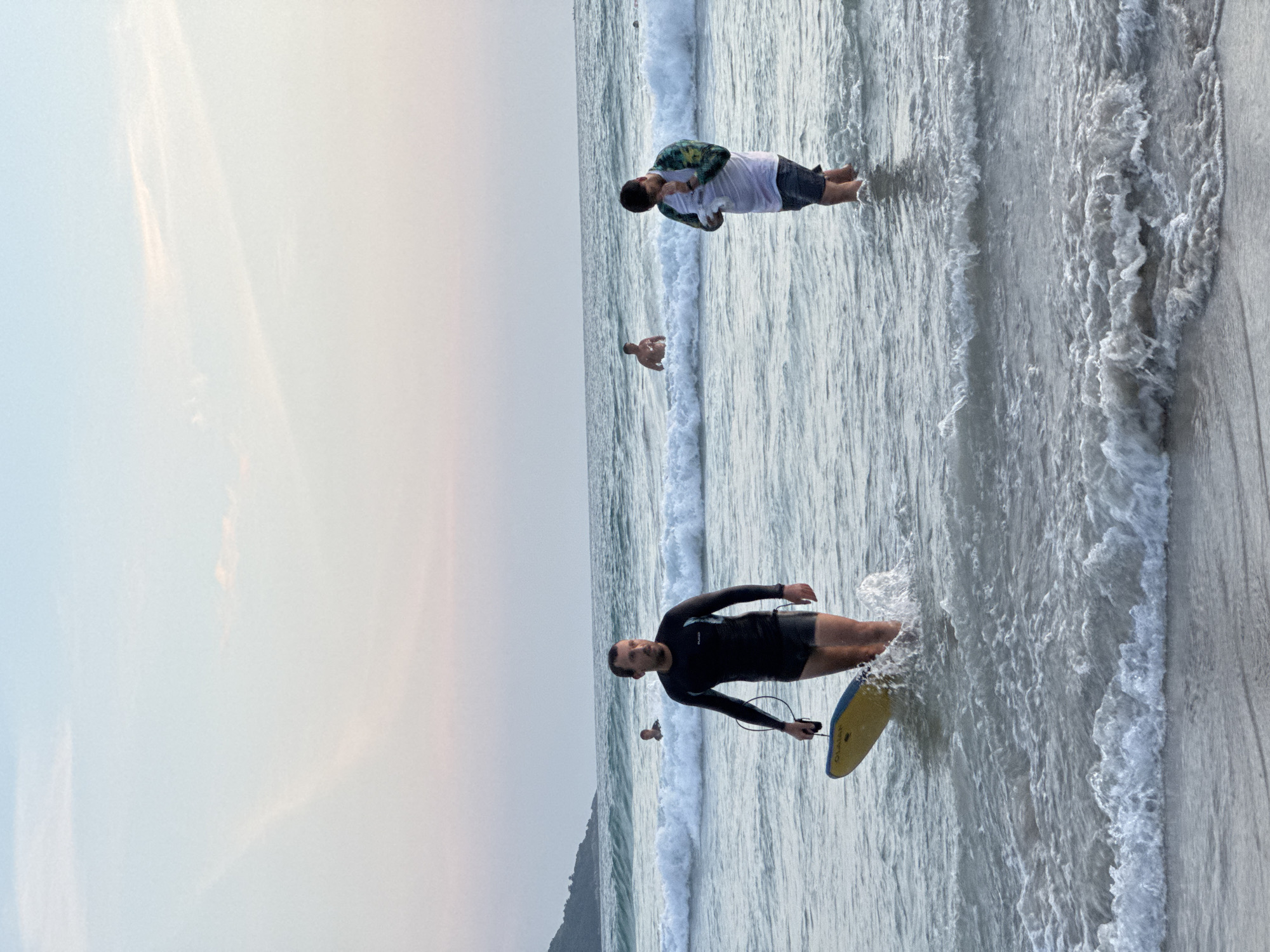 In Karon, Thailand, at dusk, a man wearing a black wetsuit and dark swim trunks walks out of the ocean in shallow water, carrying a yellow boogie board. He is partially submerged and kicking up water. To his right, another man stands in the water, wearing a white t-shirt with a floral pattern and dark swim trunks, holding a white object to his face. In the background, a man in swim trunks is visible in the ocean, and beyond him, small waves break on the shore. The sky is a pale gradient of pink and blue, with wispy clouds. A distant coastline is visible on the left horizon.