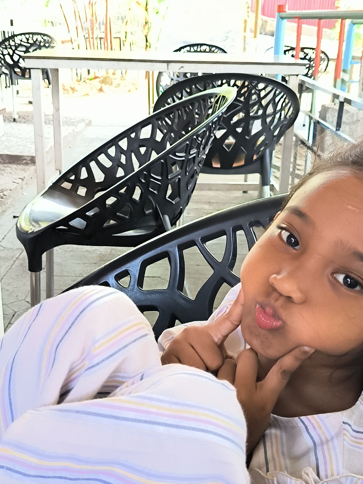 A young child with dark skin and eyes is sitting in a black chair with a decorative open-weave design. She is wearing a light-colored striped shirt or pajamas and is making a "duck face" expression while holding her cheeks with her fingers. Several similar black chairs are stacked behind her, sitting under white tables. The background shows an outdoor patio area with elements of green foliage and what appears to be a simple structure with red and blue accents. The image was captured in Matang, Indonesia.