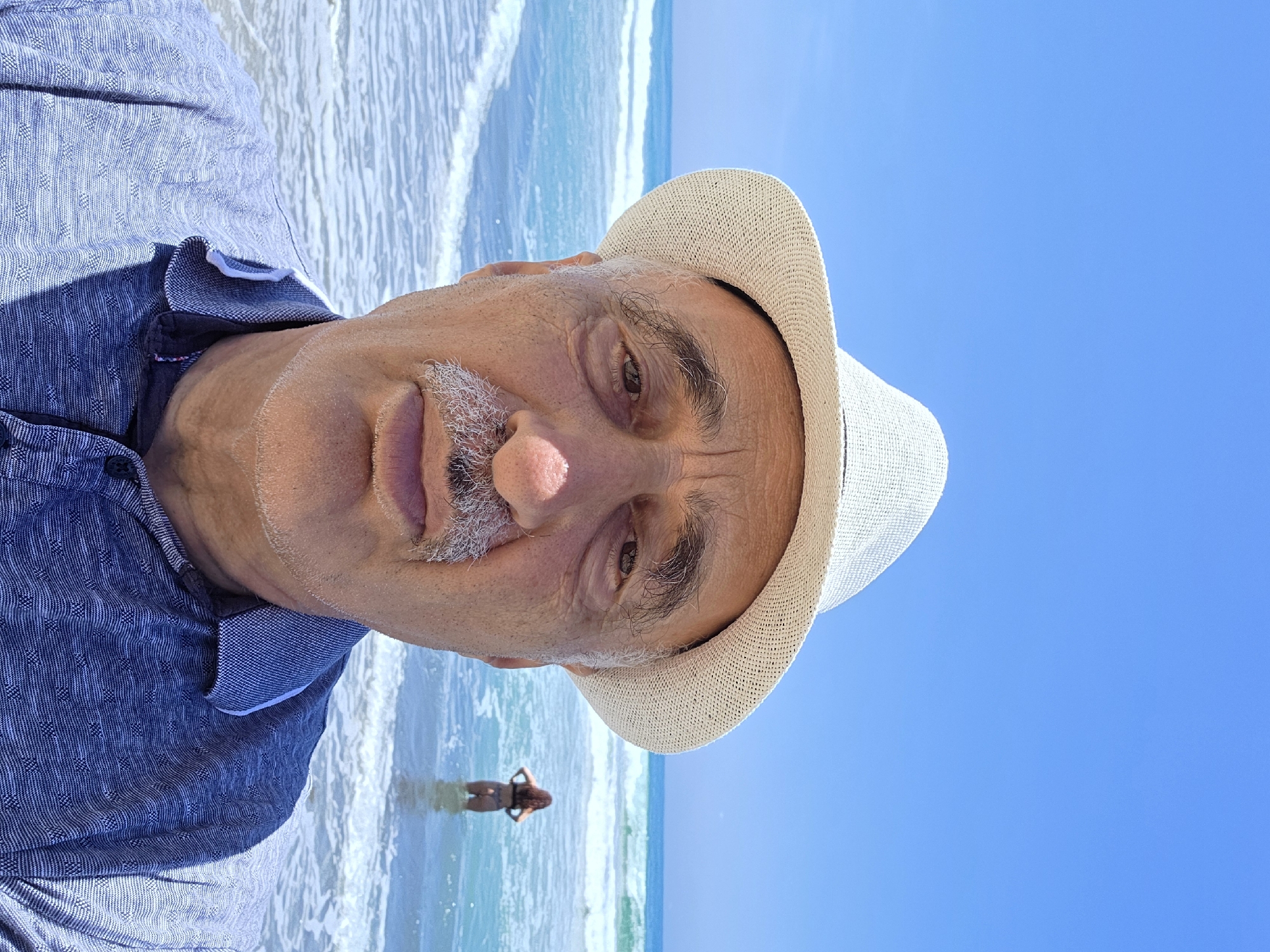 A man wearing a white fedora hat and a blue patterned collared shirt is taking a selfie at the beach. He is facing the camera directly, with the ocean and a clear blue sky in the background. A woman in a bikini is visible in the water behind him, facing away from the camera. The waves are gently rolling in. The man has a mustache and appears to be middle-aged or older. The scene is set at the beach in San Clemente, California, United States of America.