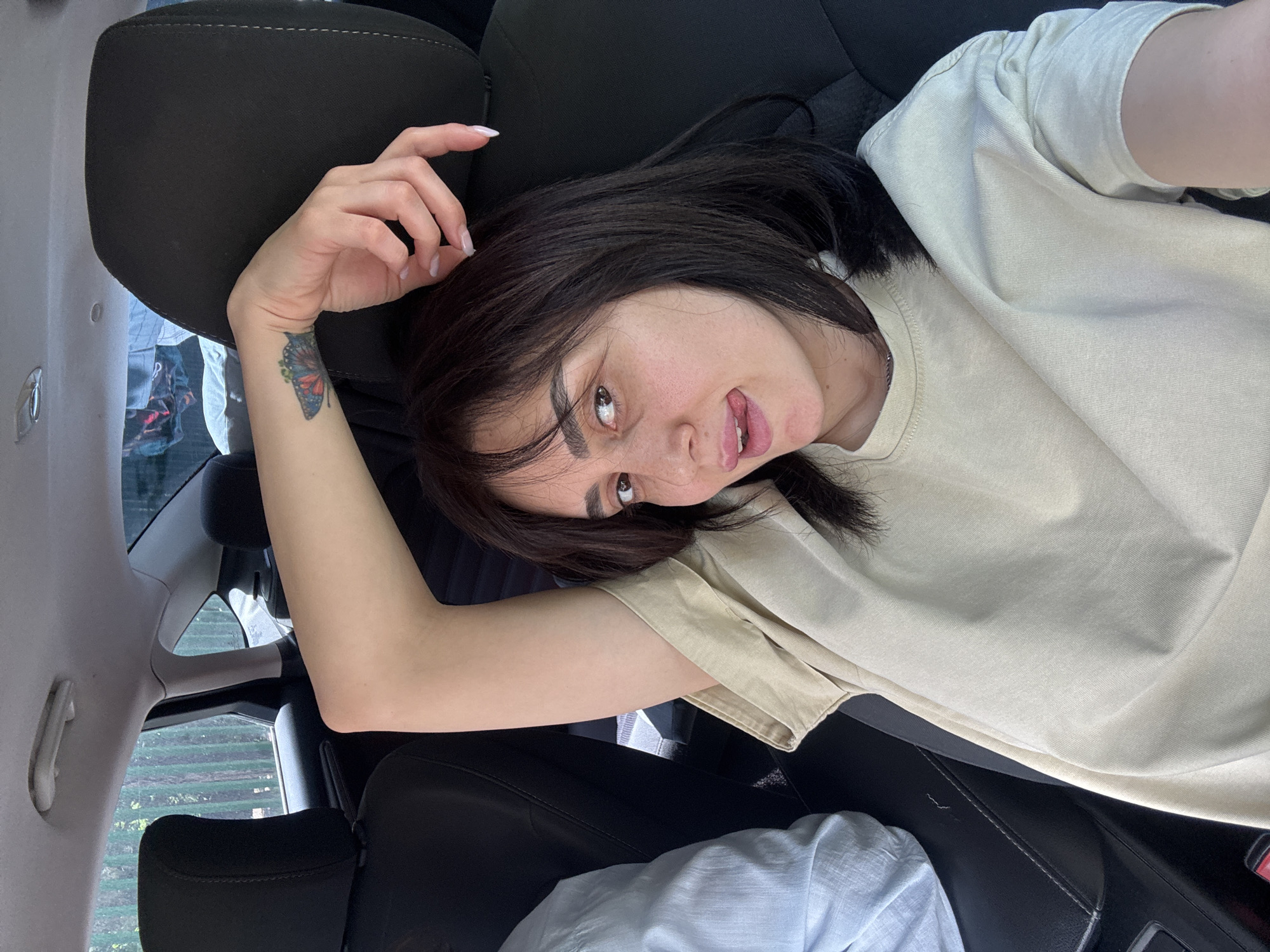 A woman is lying in the passenger seat of a car. She has dark hair, fair skin, and a butterfly tattoo on her left wrist. She is wearing a beige t-shirt. The car interior consists of black seats and a window showing greenery outside. The location is likely Tbilisi, Georgia.