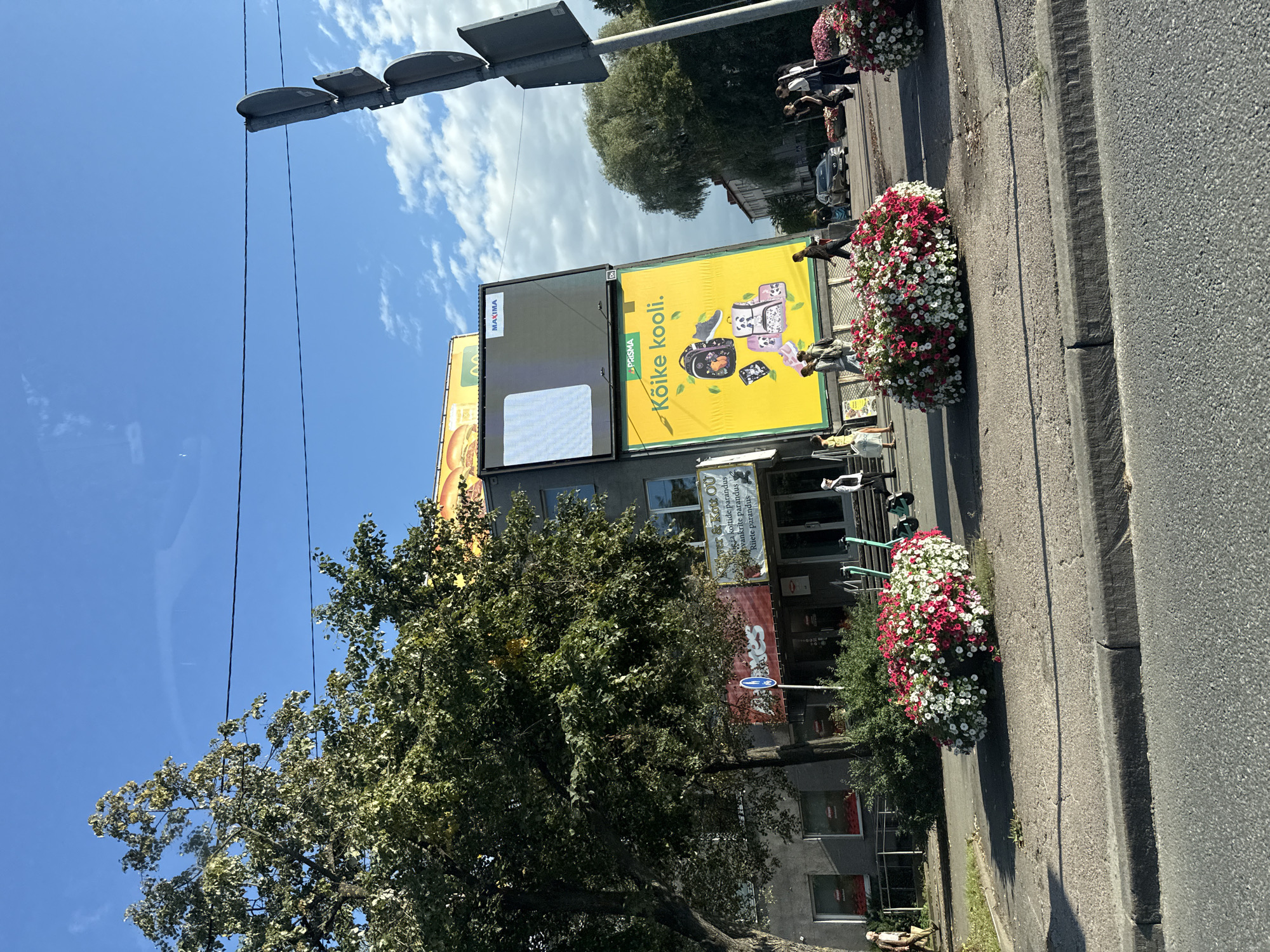 The image depicts a street scene in Tallinn, Estonia, on a sunny day. In the foreground, a grey asphalt road is visible with a concrete curb. Two large, circular flower beds filled with red and white petunias are placed along the sidewalk. A paved sidewalk runs parallel to the road, with a group of trees to the left and a building with advertisements to the right.
The building is a commercial establishment with a dark facade. A large, bright yellow billboard with the text "PRISMA Kõike kooli." and images of backpacks and a shoe is prominently displayed. Above this, a large digital screen shows a blue and white pattern, and a smaller sign reads "MAXIMA." Another advertisement featuring a hamburger is partially visible at the top.
Several people are walking on the sidewalk. Some are individuals, while others are in small groups. Two teal electric scooters are parked near the entrance of the building. Traffic lights are visible on a pole on the right side of the image. In the background, a few cars are driving on the street, and more trees and buildings can be seen. The sky is blue with scattered white clouds. The overall atmosphere is that of an urban commercial area on a clear day.