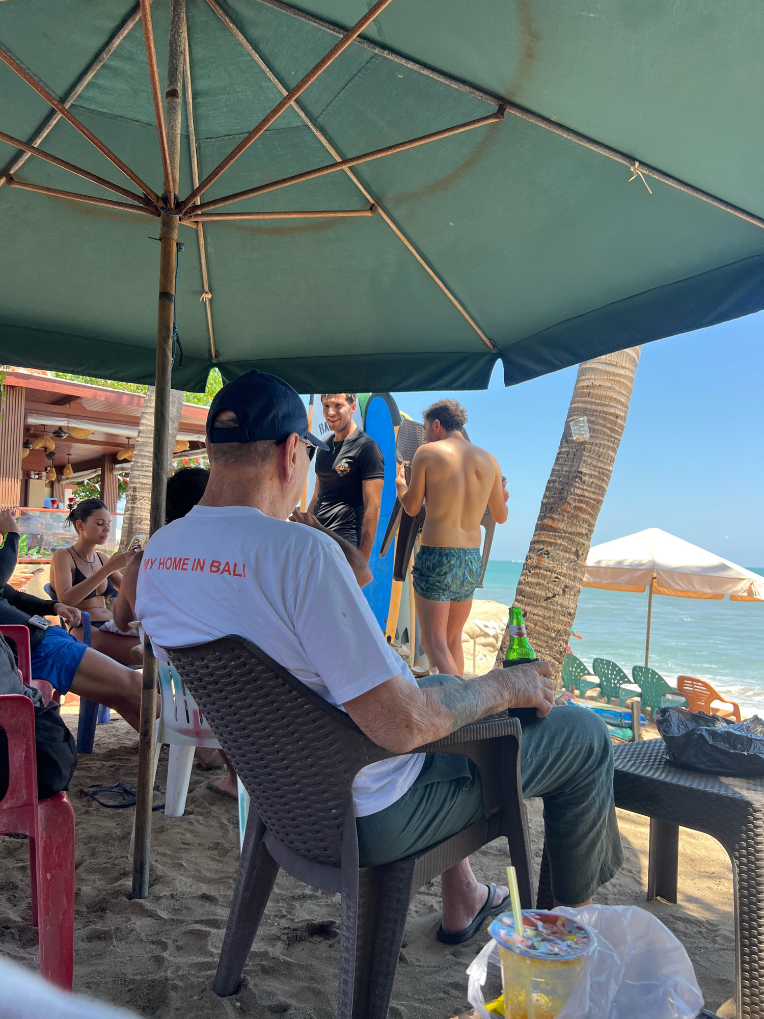 An outdoor beach scene in Kuta, Indonesia, depicts a group of people relaxing under a large green umbrella. In the foreground, a person wearing a dark blue baseball cap and a white t-shirt with red text reading "MY HOME IN BALI" sits in a brown plastic chair, holding a green bottle with a beverage. To their right, a table with a black plastic bag rests on the sand. Further back, another individual in a black t-shirt stands and smiles, while a shirtless person in patterned swim trunks is seen from behind, holding a chair. Several surfboards are visible leaning against a palm tree. The beach is sandy, with the ocean and a clear blue sky in the background. In the distance, beach chairs and a white umbrella are set up. The overall atmosphere suggests a casual leisure activity at a beachfront establishment.