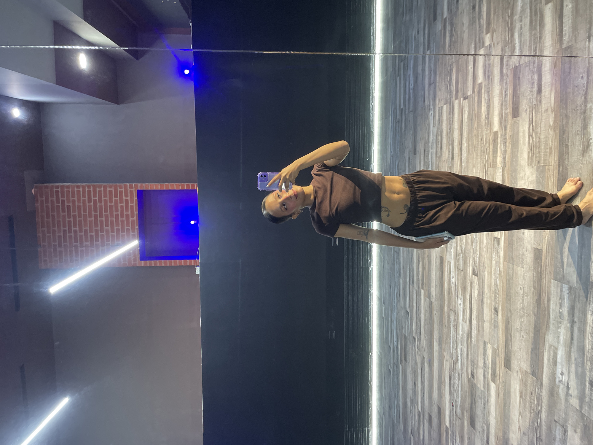 A full-shot, low-angle, full-body selfie of a woman in a dark brown crop top and loose-fitting dark brown sweatpants is taken in a dance studio. The woman is standing with her left foot forward and her right foot behind, holding a purple phone in her right hand to take the picture. She has a lean build and visible tattoos on her torso and left arm. Her hair is pulled back into a bun, and she is wearing subtle makeup. The background features a dark, reflective wall, a wooden floor with light-colored planks, and a brick-patterned wall with blue lighting. The setting is likely a dance studio in Pampatar, Venezuela, suggested by the context of the request.