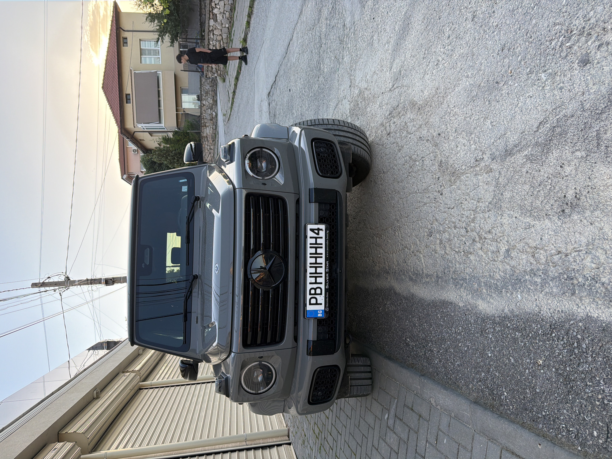 The image shows a gray Mercedes-Benz G-Class SUV parked on a concrete street in Asenovgrad, Bulgaria. The vehicle's front is facing the viewer, with its license plate reading "PBHHHHH4". In the background, there is a residential building with a beige facade and a red tiled roof. A person wearing dark clothing is walking on the street near the building. Utility poles with wires are visible in the upper left portion of the image. A building with a roll up garage door is located on the left side of the image. The lighting indicates the photo was taken during the day.