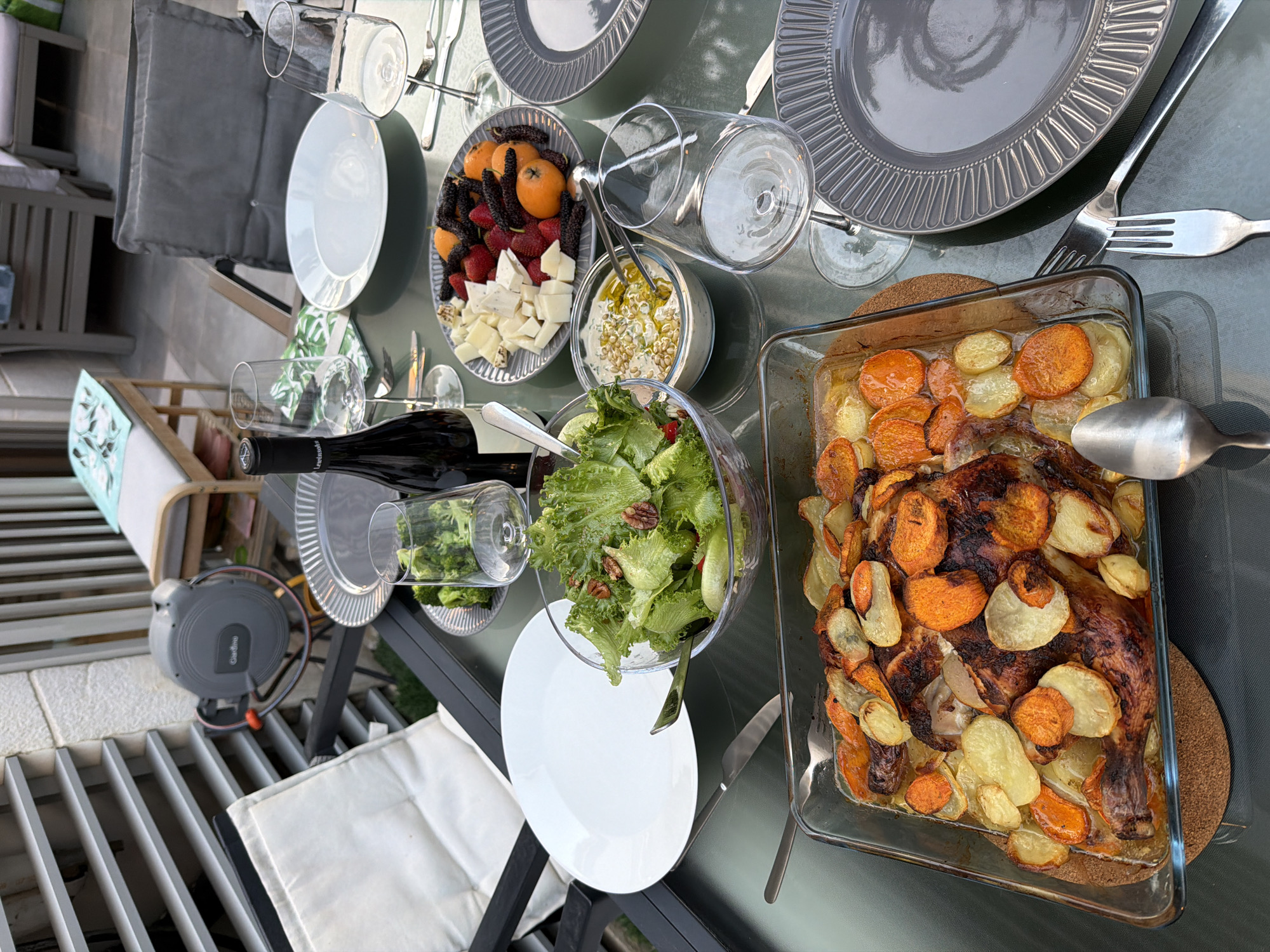 The image shows a dining table laden with food, set up outdoors. The table is covered with a dark green cloth, possibly on a patio. The setting appears to be in Herzliya, Israel, based on the architectural style and the presence of a Gardena hose reel, a common gardening brand.
Food items include:
- Roast chicken with potatoes and carrots in a glass baking dish
- A salad with green lettuce and other ingredients in a glass bowl
- A plate with mixed fruit and cheese, including strawberries, mulberries, apricots, and cheese cubes
- A bowl of dip, possibly hummus or baba ghanoush, with herbs and pine nuts
- A small plate of broccoli
Table settings include:
- Gray patterned plates
- Wine glasses, some filled with a beverage and one with broccoli
- Silverware: forks, knives, and spoons
Other details:
- A bottle of wine is lying on the table
- White napkins are placed near the table's edge
- A gray outdoor chair is visible in the background
- A multi-level, open-slat patio surface is visible with the Gardena hose reel
- A small, decorative wooden stand with a teal-colored, leaf-patterned covering stands to the side.
The overall impression is a prepared meal ready to be served on an outdoor patio. No people are directly visible in the image.