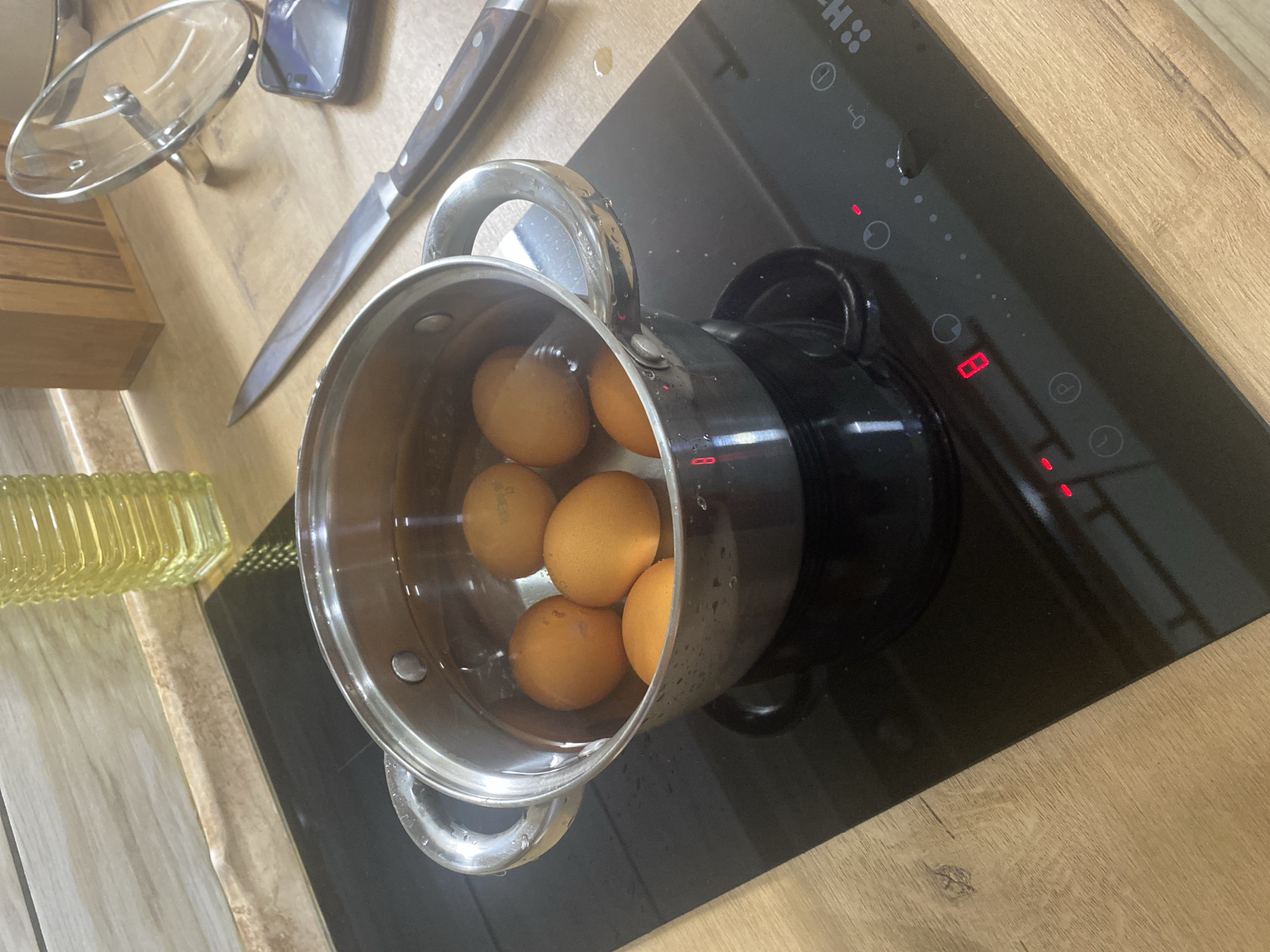 This image captures a domestic kitchen scene in the city of Копнино, Russia. A stainless steel saucepan containing six brown eggs submerged in water is positioned on the front-left burner of a black induction stovetop. The stovetop's integrated control panel, located to the right of the pot, displays the number '8' in red LED, indicating the heat level, alongside various touch-sensitive icons for power, timer, and a child lock, and a partial "HA..." branding. Small water droplets are visible on the stovetop surface and the exterior of the saucepan, suggesting active boiling or condensation.
The countertop, made of a light brown wood-grain laminate, extends across the scene. To the right of the stovetop, a long chef's knife with a dark wooden handle and a silver blade rests horizontally on the countertop, pointing left. Further to the right, a clear glass pot lid with a silver knob is propped upright, leaning against an unseen object or the wall. A dark-colored smartphone lies flat on the countertop adjacent to the lid. In the background, a light-toned, wood-grained backsplash is visible, above which sits a tall, yellow, ribbed glass bottle, possibly an oil dispenser. A wooden block, potentially a knife holder, is also partially visible behind the lid and smartphone.