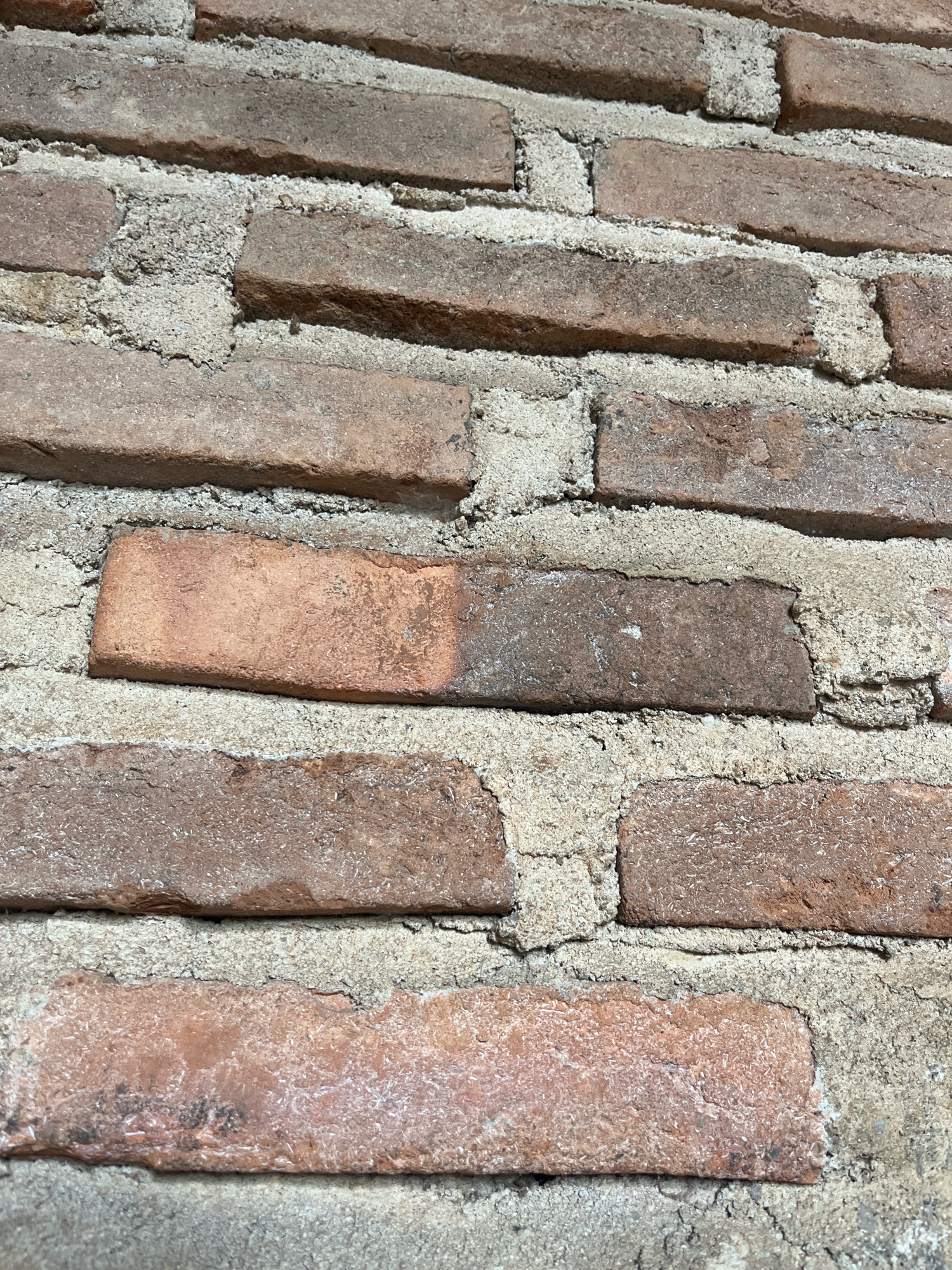 The image shows a close-up view of an old brick wall. The bricks are rectangular and appear to be made of fired clay, exhibiting a range of reddish-brown hues with some darker patches and signs of wear. They are laid in a staggered pattern, with rough, light-colored mortar filling the gaps between them. The mortar is textured and shows some cracks and crumbling. The lighting suggests the image was taken outdoors, with shadows indicating a directional light source, possibly sunlight. There are no people or other objects visible in the frame, focusing solely on the texture and material of the wall. The context of Sidomulyo, Indonesia, suggests this could be a historical building, a section of a boundary wall, or an architectural feature in that region.