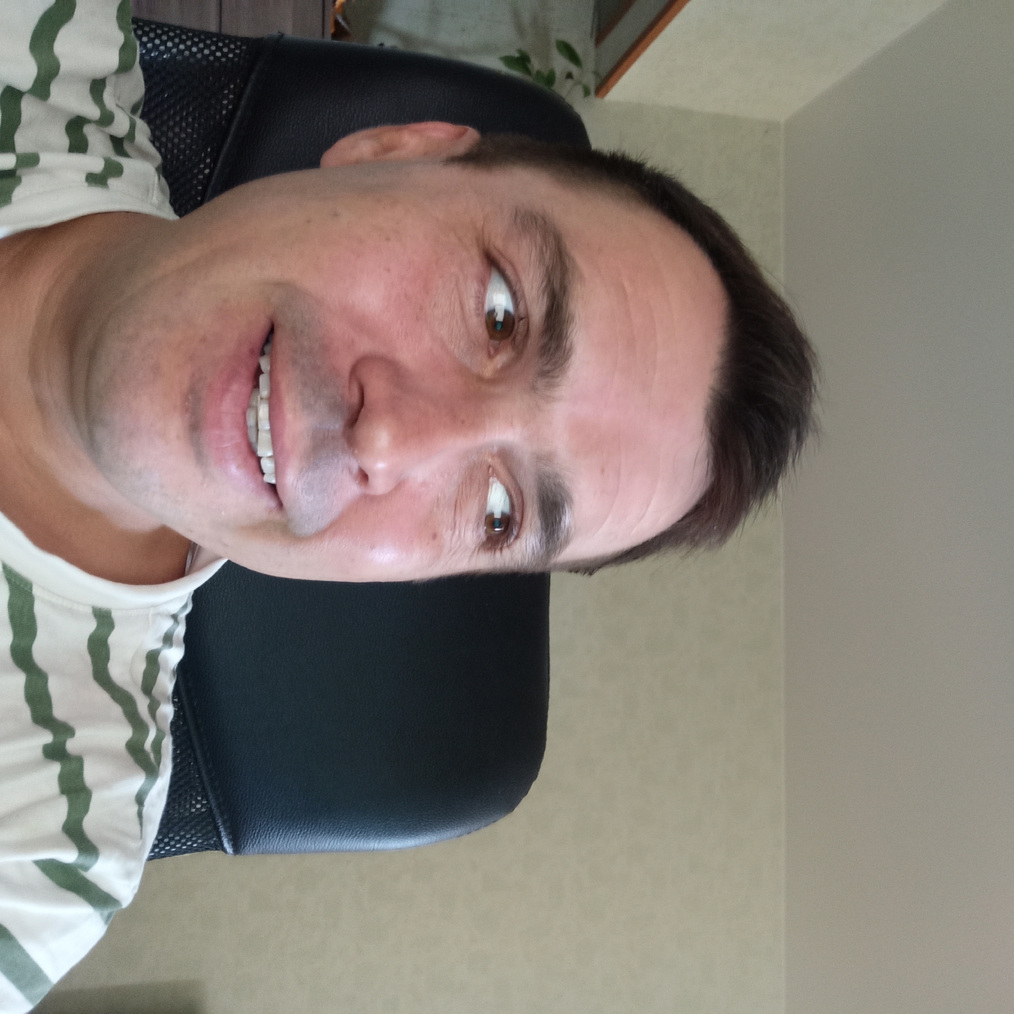 A man with short, dark brown hair is sitting in a black office chair, looking to his right with a slight smile. He is wearing a white t-shirt with green horizontal stripes. The background appears to be an indoor setting with a light-colored wall and a shelf with some greenery visible on the left. The overall lighting suggests it is daytime. The image is a close-up selfie, focusing on the man's face and upper body. No specific location in Nizhny Novgorod, Russia, is identifiable from the visual information presented in the image.