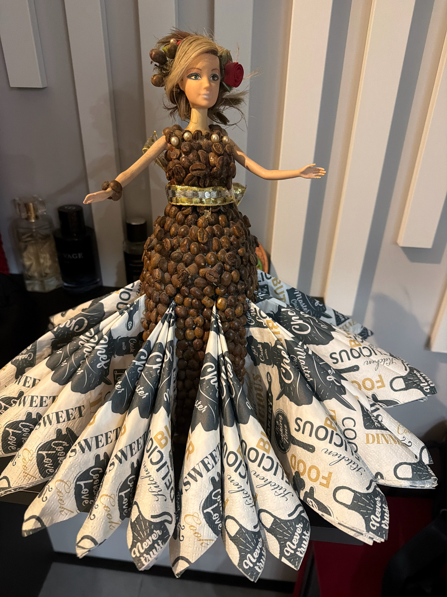 A doll is displayed in a white, ribbed environment. The doll has blonde hair with a red flower and brown accessories. Her dress is made of coffee beans and features a gold belt with pearl accents. The doll's skirt is made of patterned napkins with text like "SWEET," "DELICIOUS," "FOOD," "KITCHEN," and "COOK." The background includes two bottles of perfume on a black surface. The location is Vniissok, Russia.