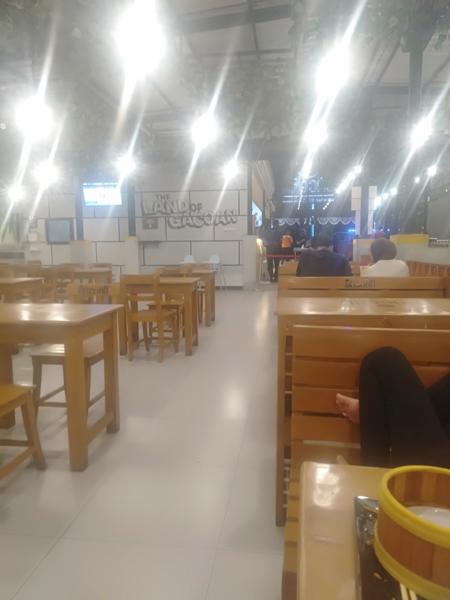 The image captures an interior scene of a restaurant or dining establishment. The environment features numerous wooden tables and chairs, arranged in a spacious layout. The lighting is bright, with multiple light fixtures visible overhead, some emitting lens flare effects. In the background, a wall displays a sign that reads "THE LAND OF GACOAN." Several patrons are present, some seated at tables, while others are partially visible. One individual is seated on a wooden bench, with their legs and feet in view. The overall impression is that of a casual, possibly busy, dining area. The image was taken in Kecamatan Pati, Indonesia.