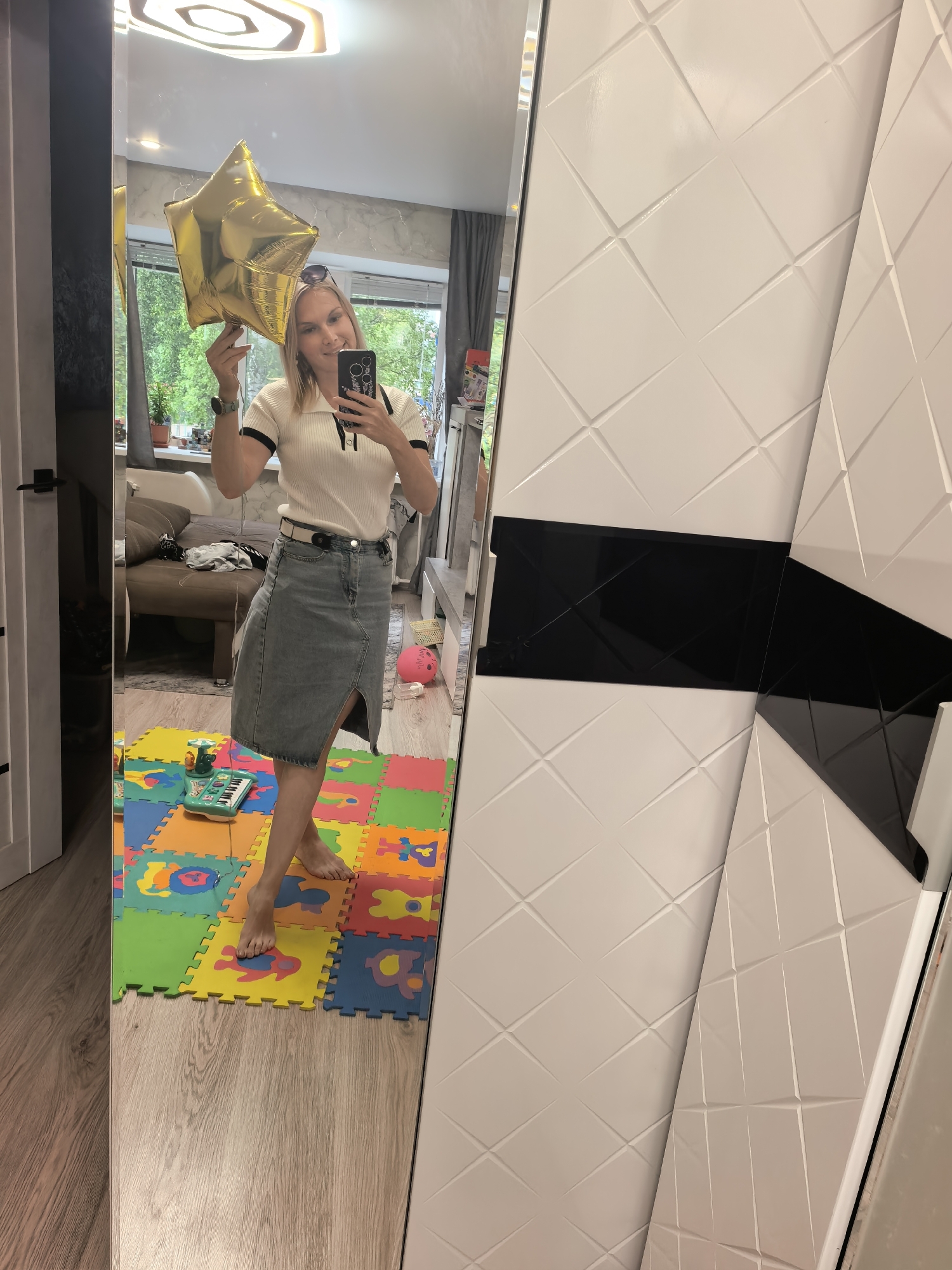 A blonde woman stands in front of a mirror, holding a gold star-shaped balloon. She is wearing a white polo shirt with black trim and a denim skirt. She is barefoot and standing on a colorful foam mat. The room appears to be a living room with a sofa and a window in the background. A closet door is visible to the left of the mirror. The right side of the image shows a white closet with a geometric pattern. The overall scene suggests an indoor setting in Sterlitamak, Russia.