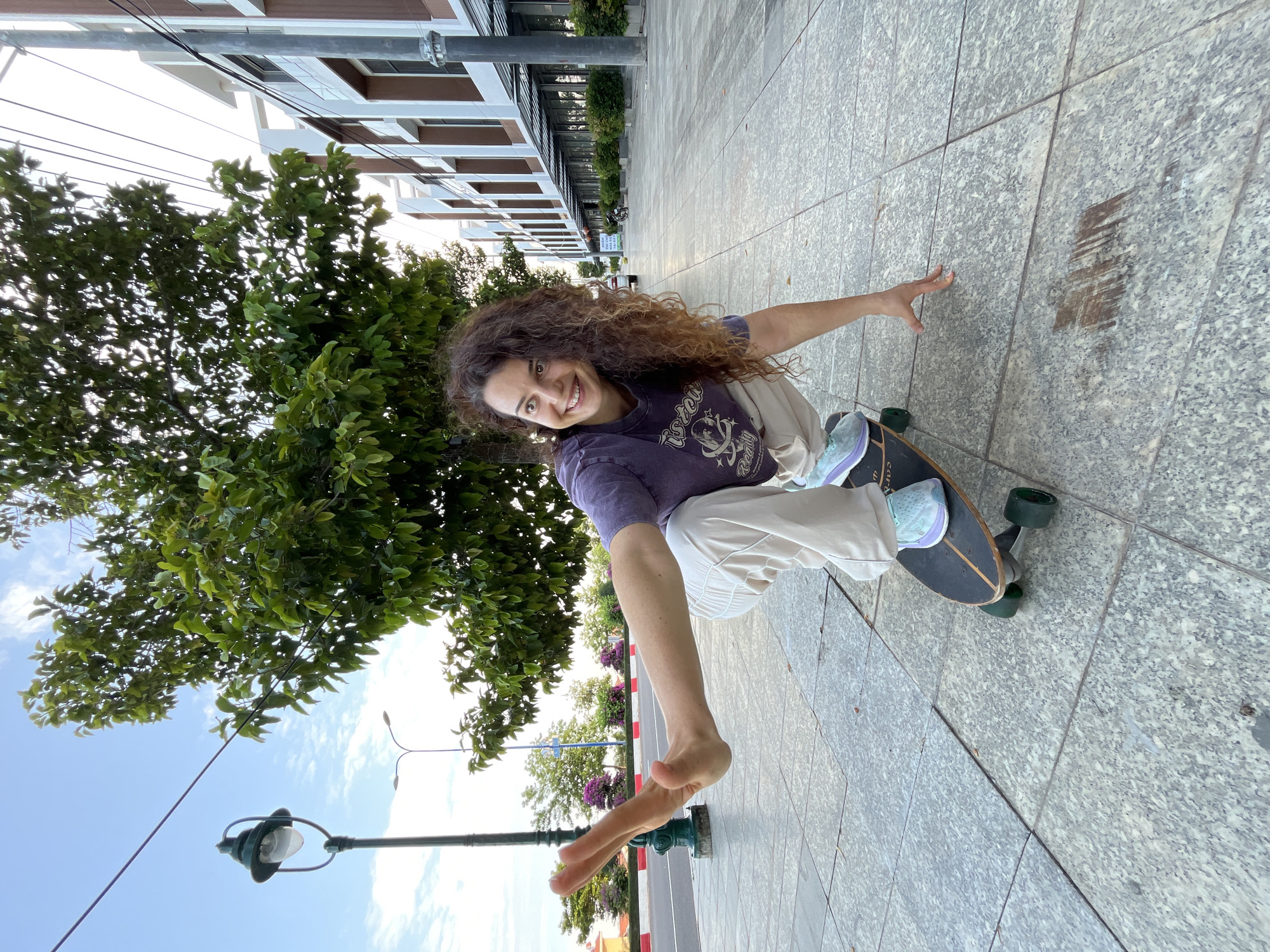 A young woman with long, curly brown hair is skateboarding on a paved sidewalk in Phu Thuy Ward, Vietnam. She is wearing a purple t-shirt and light-colored pants. She is in a crouching position on the skateboard, with her left arm extended and her right hand touching the ground for balance. The skateboard has green wheels. The sidewalk is made of large, grey stone tiles. In the background, there is a tree with green leaves, a lamppost, a street with a red and white curb, and modern buildings. The sky is blue with scattered clouds.