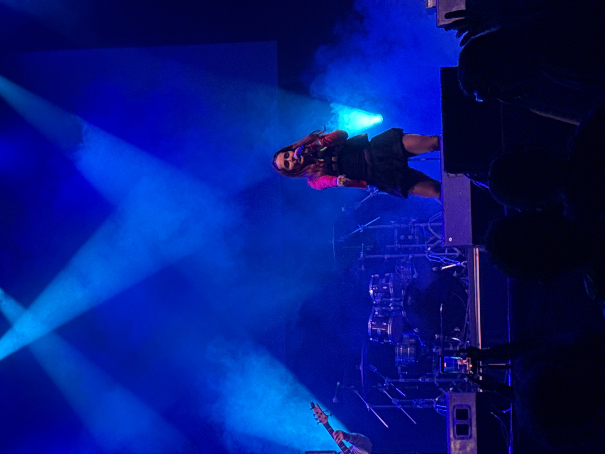A woman is singing on stage at a concert in Vilanova i la Geltrú, Spain. She is wearing a black dress with a pink jacket and holding a microphone. Blue stage lights illuminate the scene. In the background, a drum set and another band member playing guitar can be seen. Silhouetted members of the crowd are visible on the right. Some of the crowd are holding up phones to take pictures or video.
