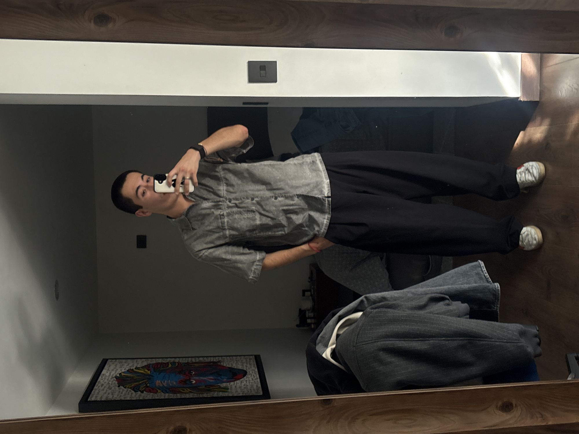 A young man is captured in a mirror selfie indoors in San Bernardino Tlaxcalancingo, Mexico. He stands facing the mirror, holding a white smartphone with a distinctive black and white case to take the photograph. He wears a casual outfit consisting of a faded grey, short-sleeved button-up shirt, black trousers, and white sneakers with light laces. A dark-strapped watch is visible on his left wrist, and he wears a ring on his left hand.
The reflection shows a simple indoor setting. A white wall is visible behind the man, featuring a dark, rectangular light switch plate positioned centrally. Bright natural light streams in from an unseen window or doorway on the upper right side of the frame, creating a highlight on the wall and floor. The floor, along with the top and bottom frames of the mirror, appears to be made of dark wood with a visible grain pattern.
In the lower left portion of the reflection, a framed piece of artwork is displayed, showing a vibrant, colorful, stylized image that resembles a face or animal, primarily in hues of red, blue, yellow, and green. Below the artwork, some unidentifiable text is present but illegible. In the lower right foreground, outside the main reflection of the room, a folded dark grey garment, possibly a pinstriped jacket or coat, rests on a surface, with another item of clothing beneath it. The overall impression is a casual, everyday moment, likely taken during the daytime due to the natural light.