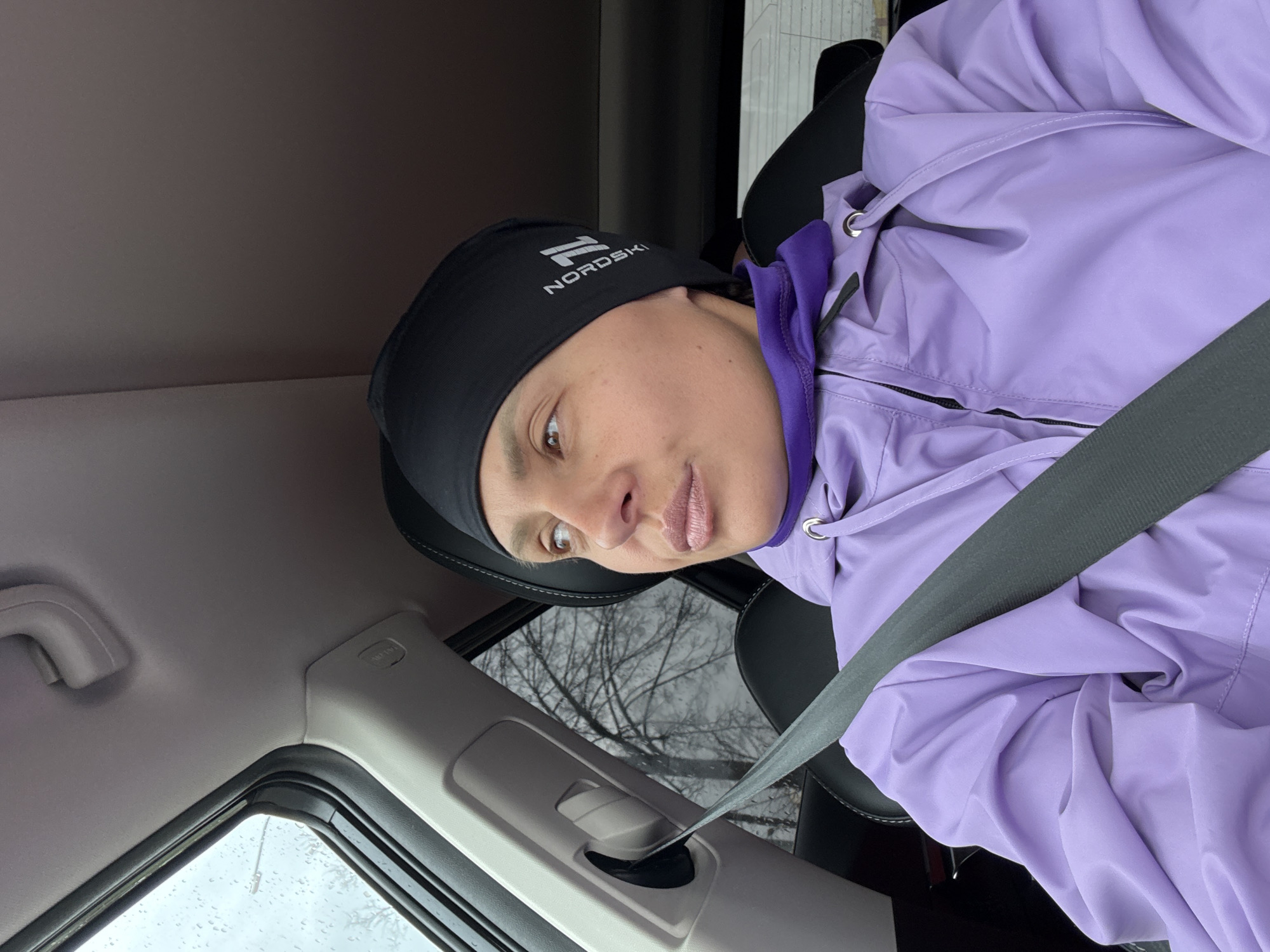 A person wearing a black beanie with the word "NORDSI" on it and a lavender jacket is sitting in a car wearing a seatbelt. The person is looking slightly down and to the left. Outside the car window, there is a view of trees with bare branches. The location is Chaikovsky, Russia.