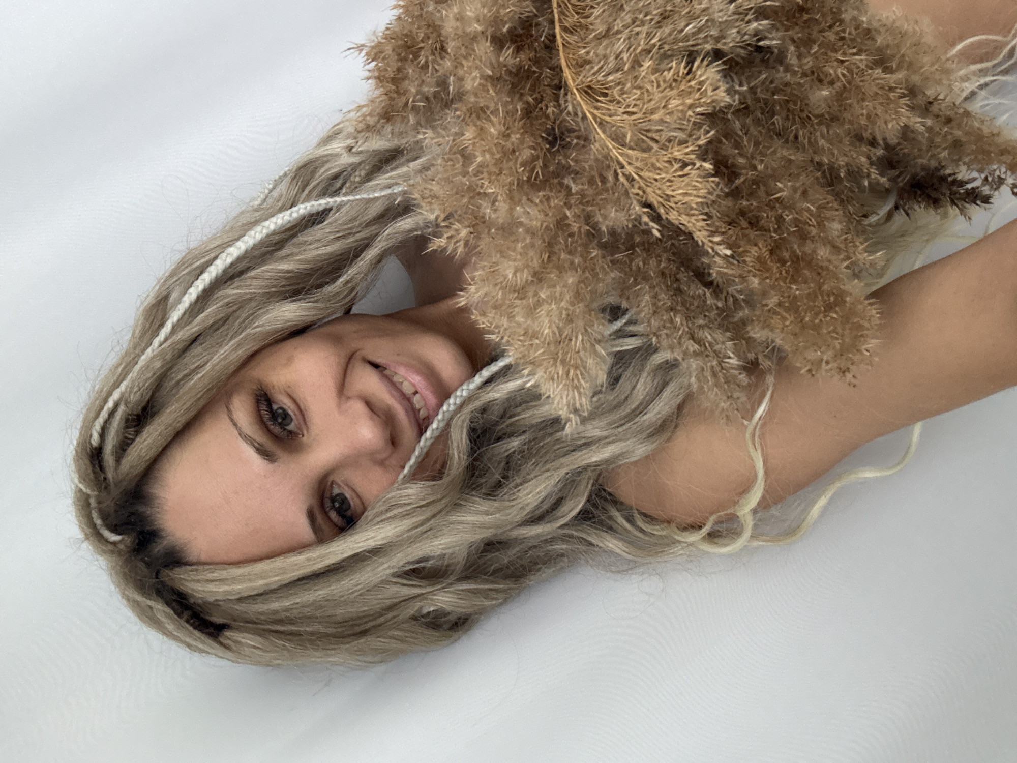 The image shows a woman with blonde hair and white braids posing with a bundle of dried pampas grass. The woman is smiling and lying on a white surface. The location is not discernable from the image and therefore cannot be linked to Rusanovo, Russia.